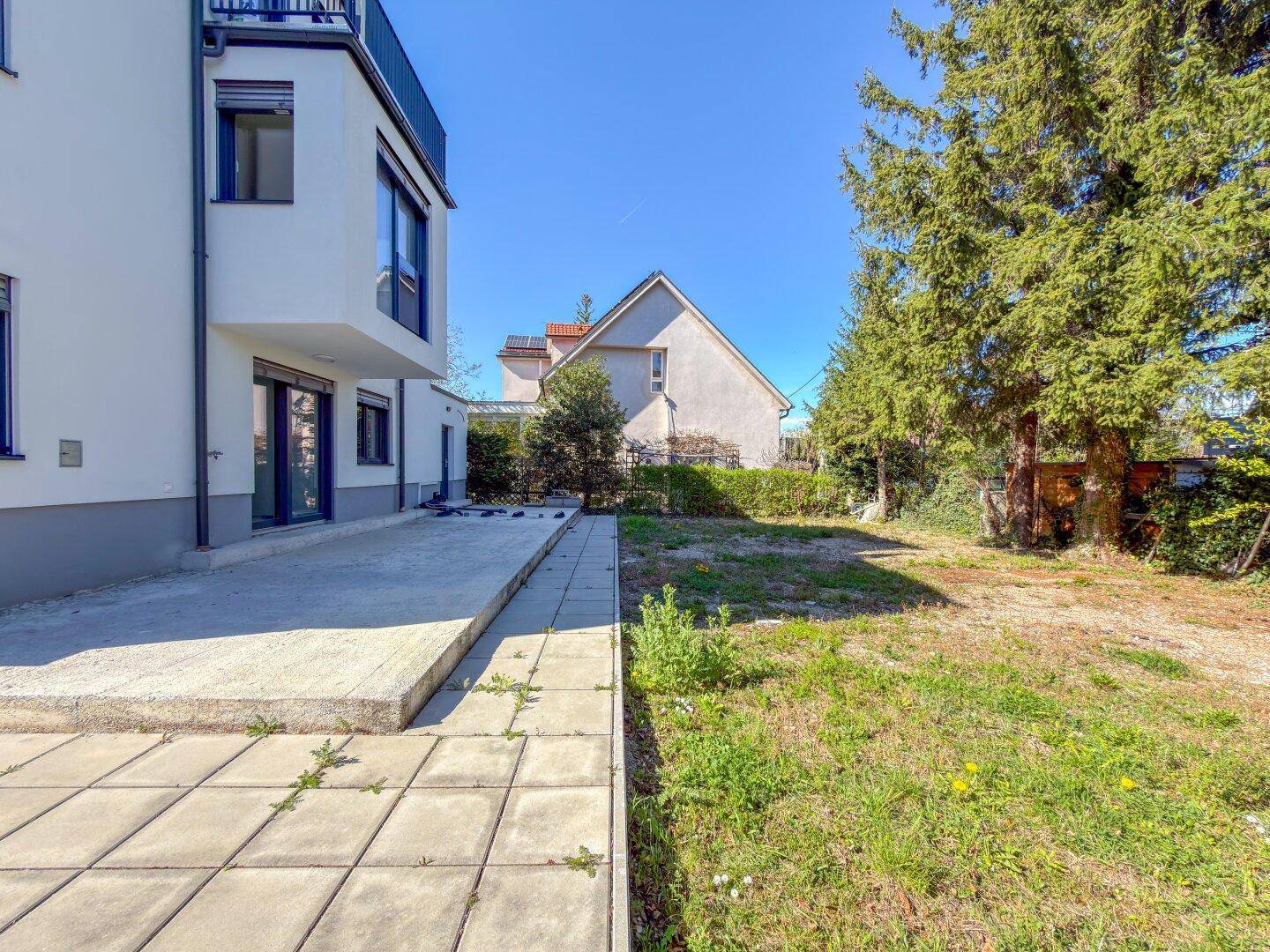Gartenbereich mit großer, gepflasterter Terrasse und altem Baumbestand neben dem modernen Gebäude.