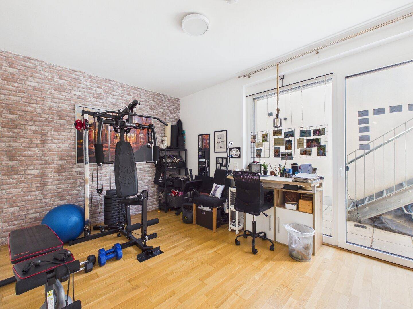 Geräumiger Raum mit Fitnessgeräten, Schreibtisch und Ziegelwandakzent.