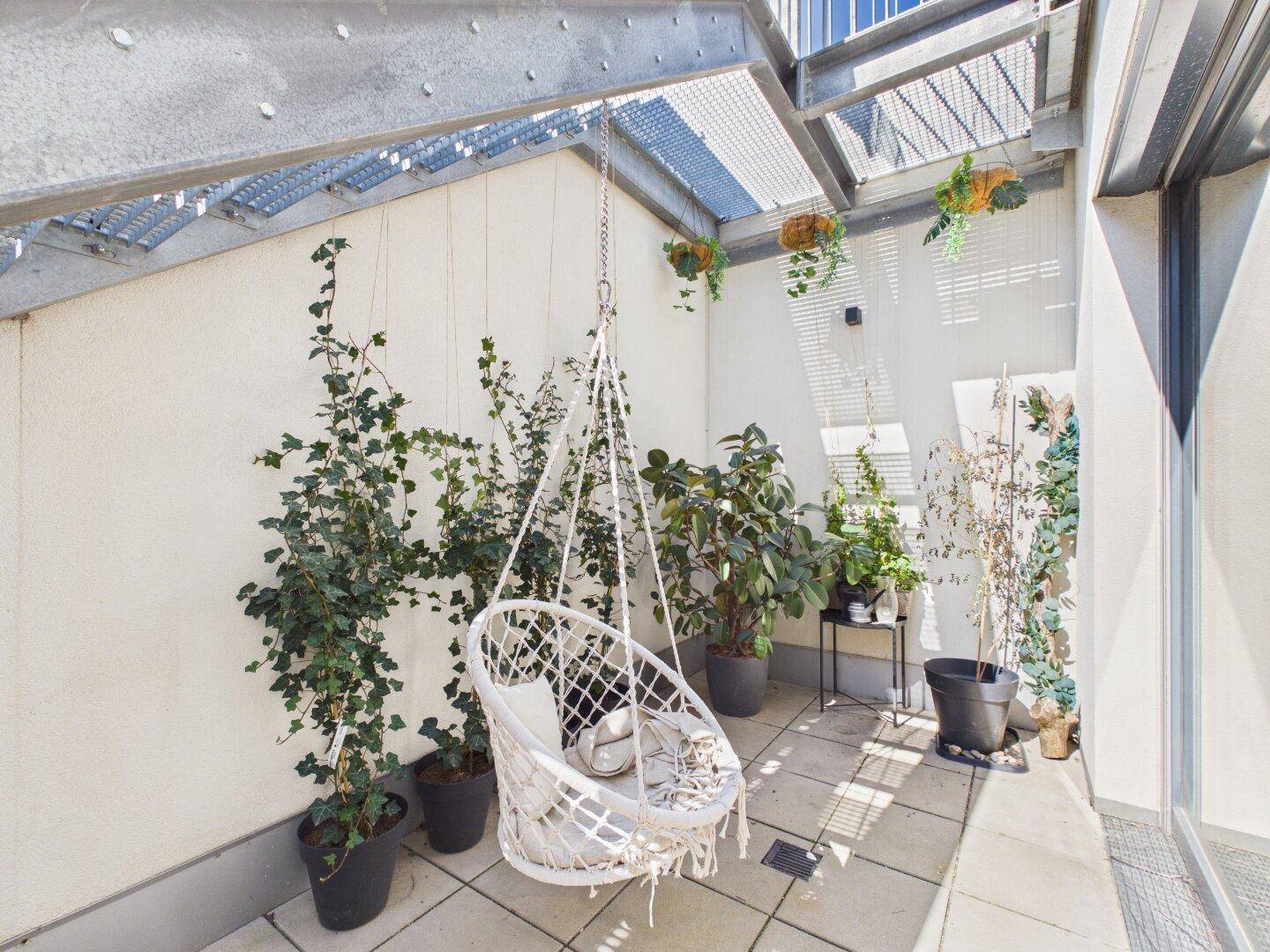 Überdachte Terrasse mit Hängesessel, vielen Pflanzen und Blick auf den Himmel.