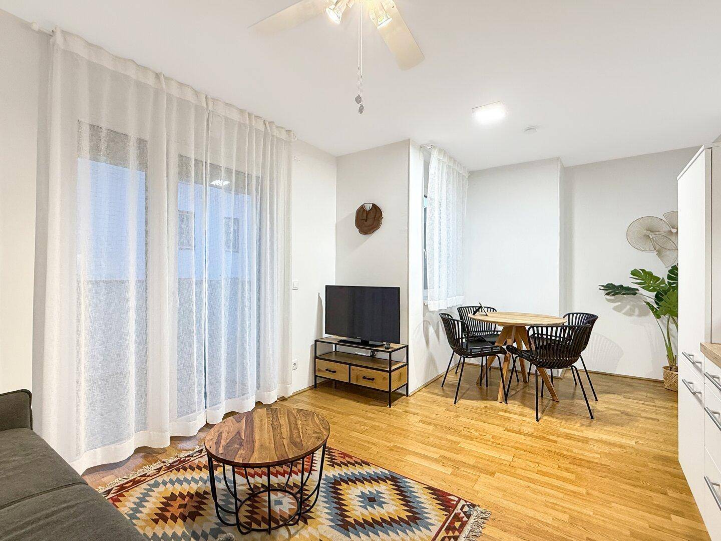 Gemütlicher Wohnbereich mit Esstisch, Sofa, Fernseher und großen Fenstern mit Vorhängen.