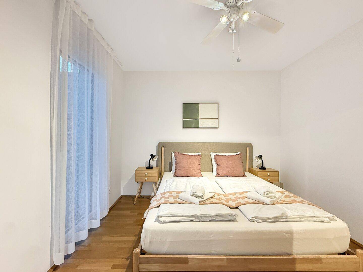 Schlafzimmer mit Doppelbett, Nachttischen, Deckenventilator und großem Fenster mit Vorhängen.