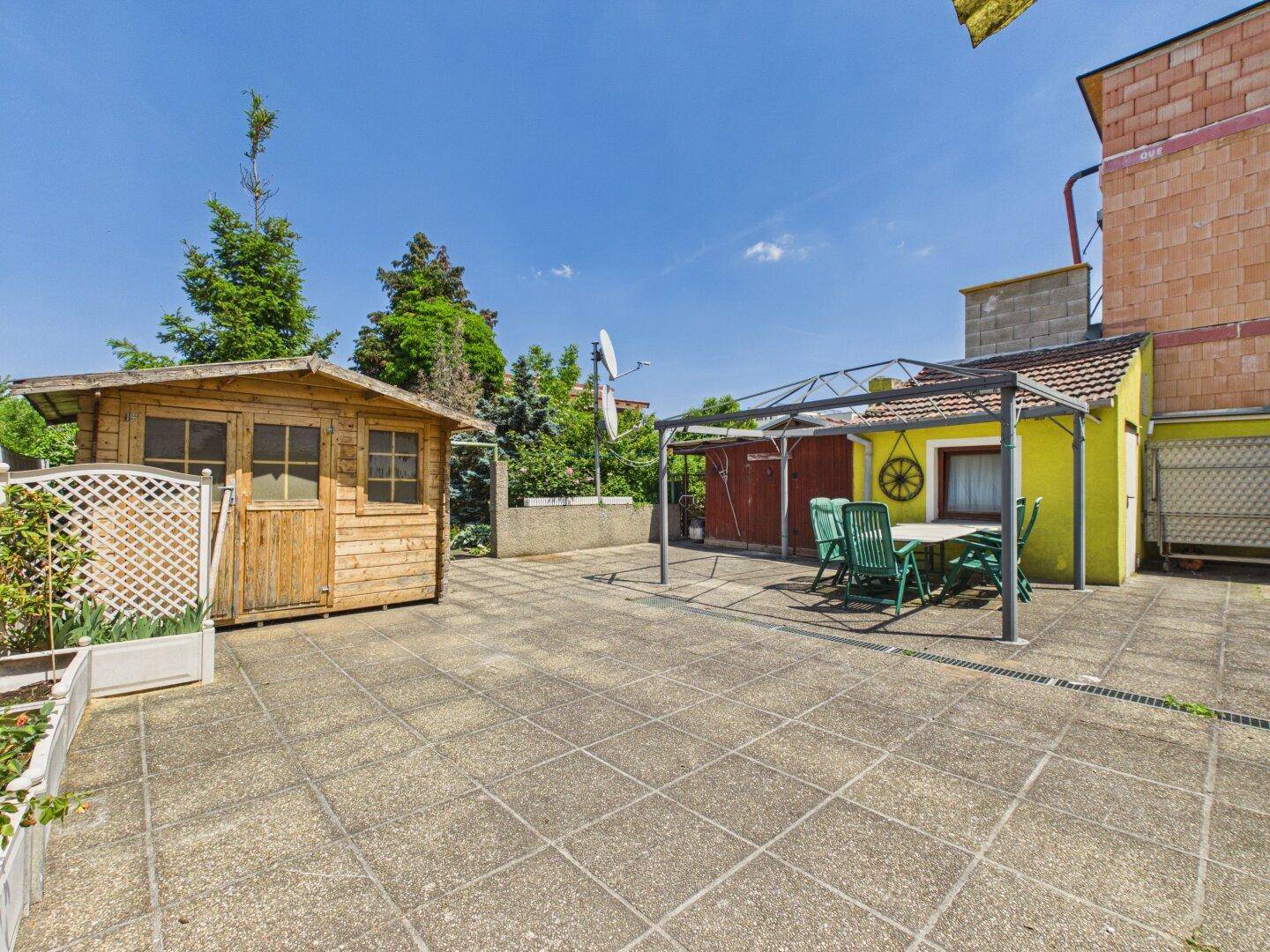 Große gepflasterte Terrasse mit Gartenhaus, Sitzgelegenheiten unter einer Pergola und sonnigem Himmel.