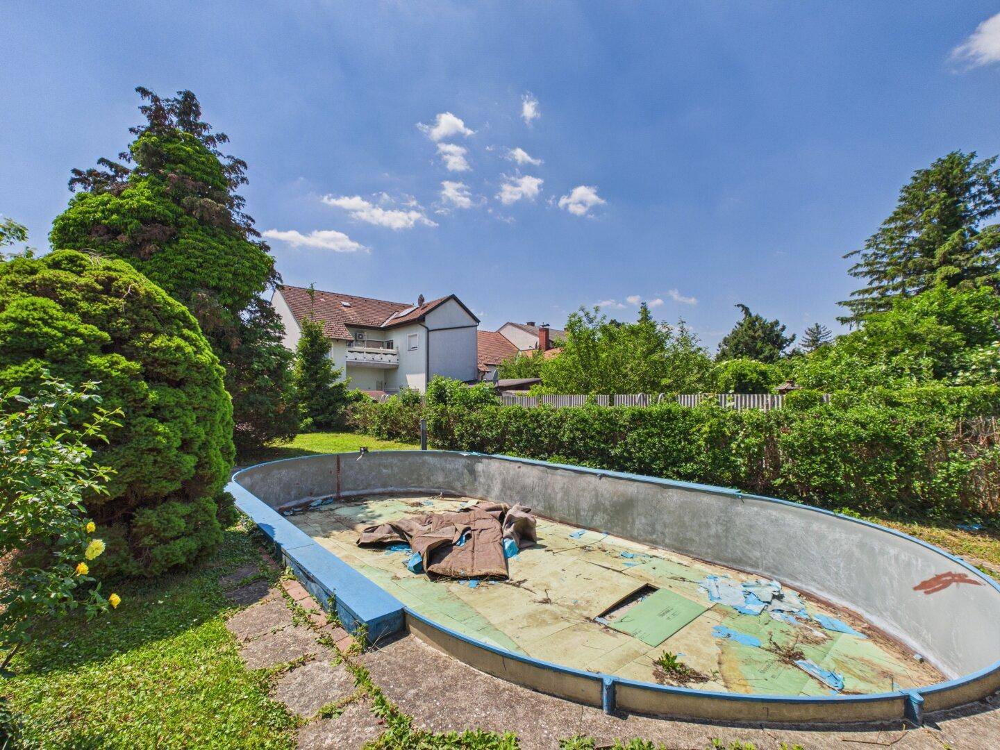 Leeres, renovierungsbedürftiges Schwimmbecken im Garten, umgeben von Bäumen und Hecken.