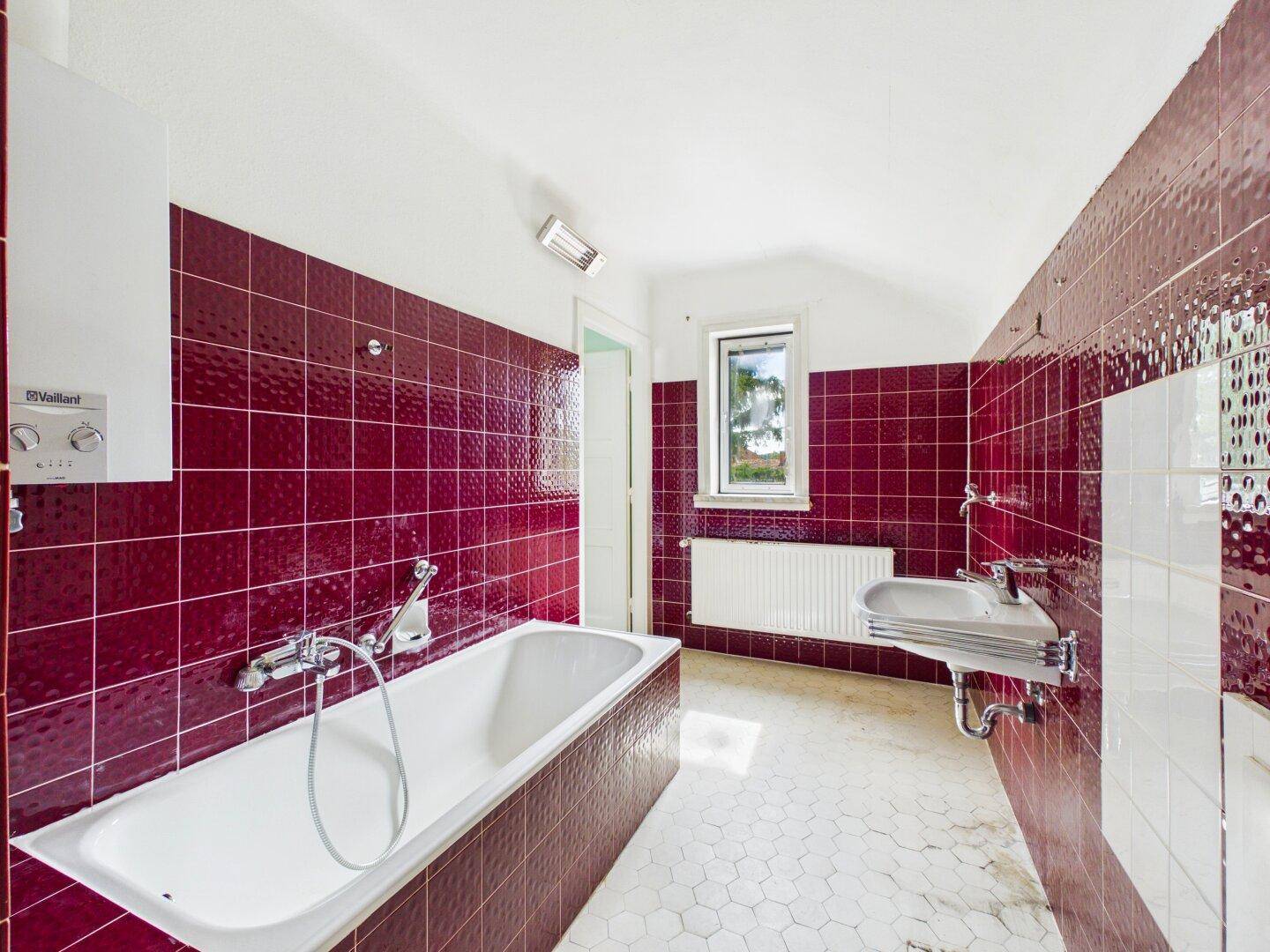 Badezimmer mit auffälligen roten Fliesen, Badewanne, Waschbecken und Fenster, benötigt Modernisierung.