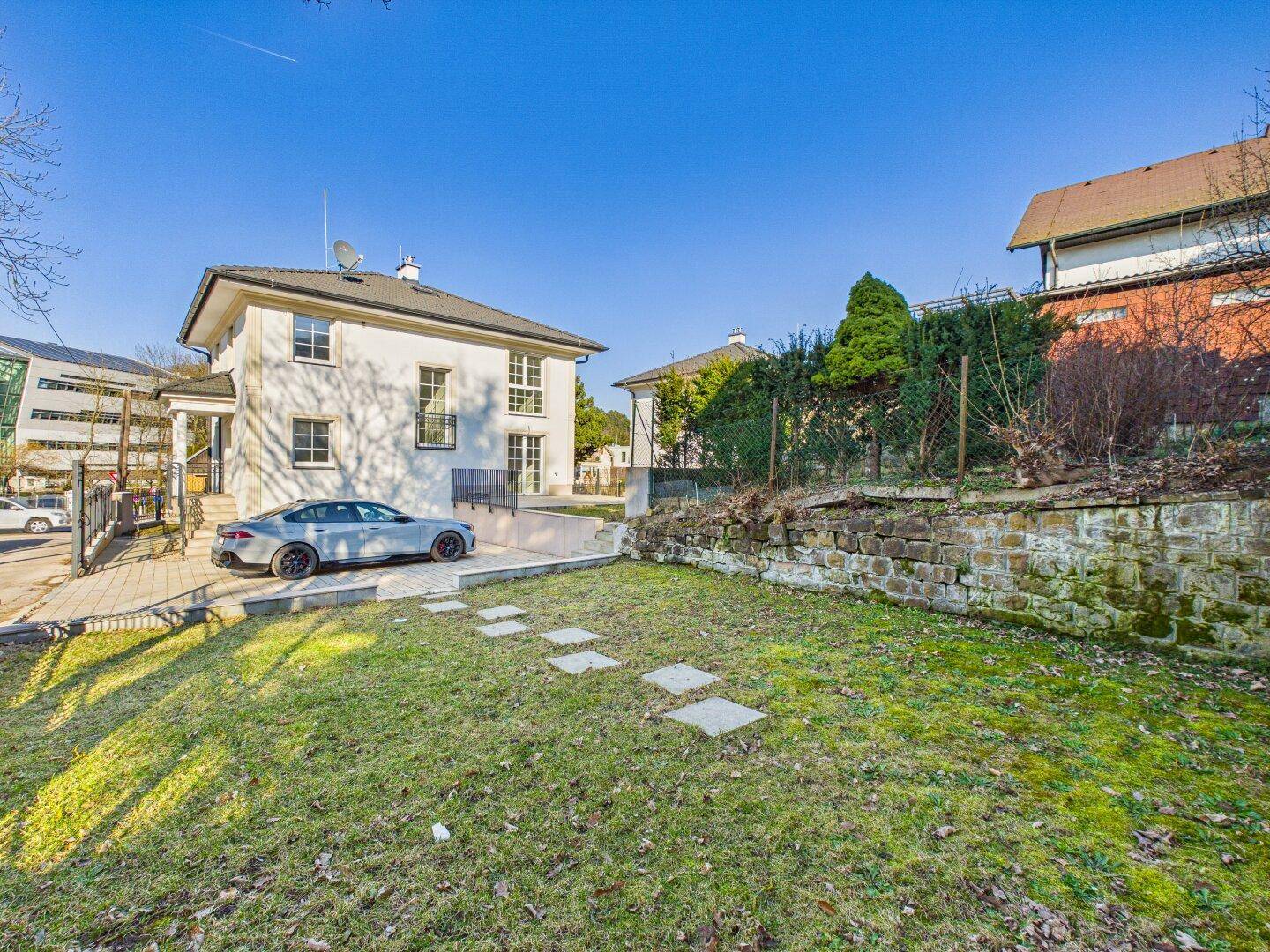 Weißes Einfamilienhaus mit gepflegtem Vorgarten, Stellplatz und einer Natursteinmauer unter blauem Himmel.
