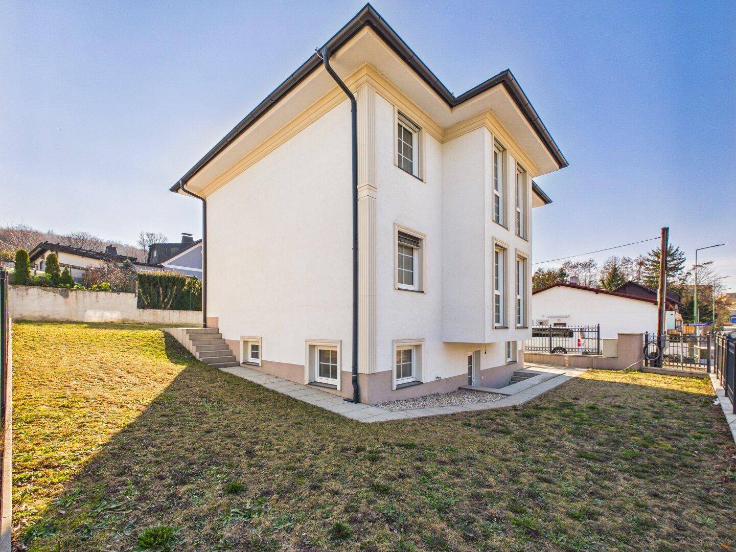 Seitenansicht des modernen Hauses mit weißer Fassade, dunklem Satteldach und abfallendem Gartenbereich.
