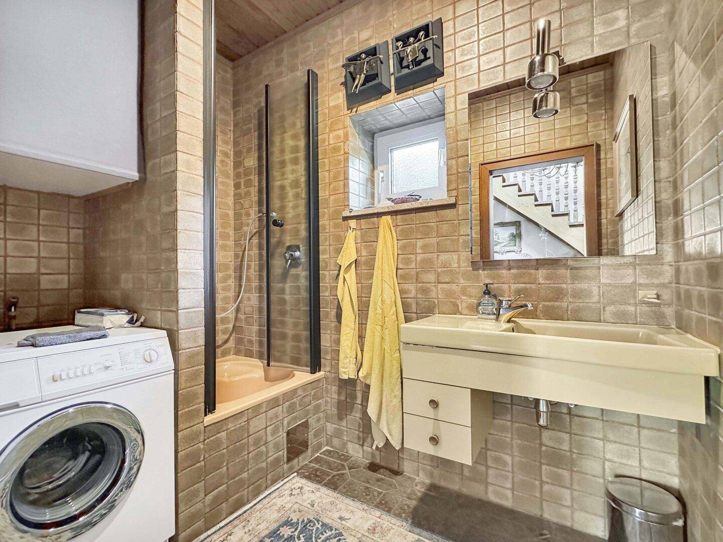 Badezimmer mit Duschkabine, Waschmaschine und braunen Fliesen im 80er-Jahre-Stil.