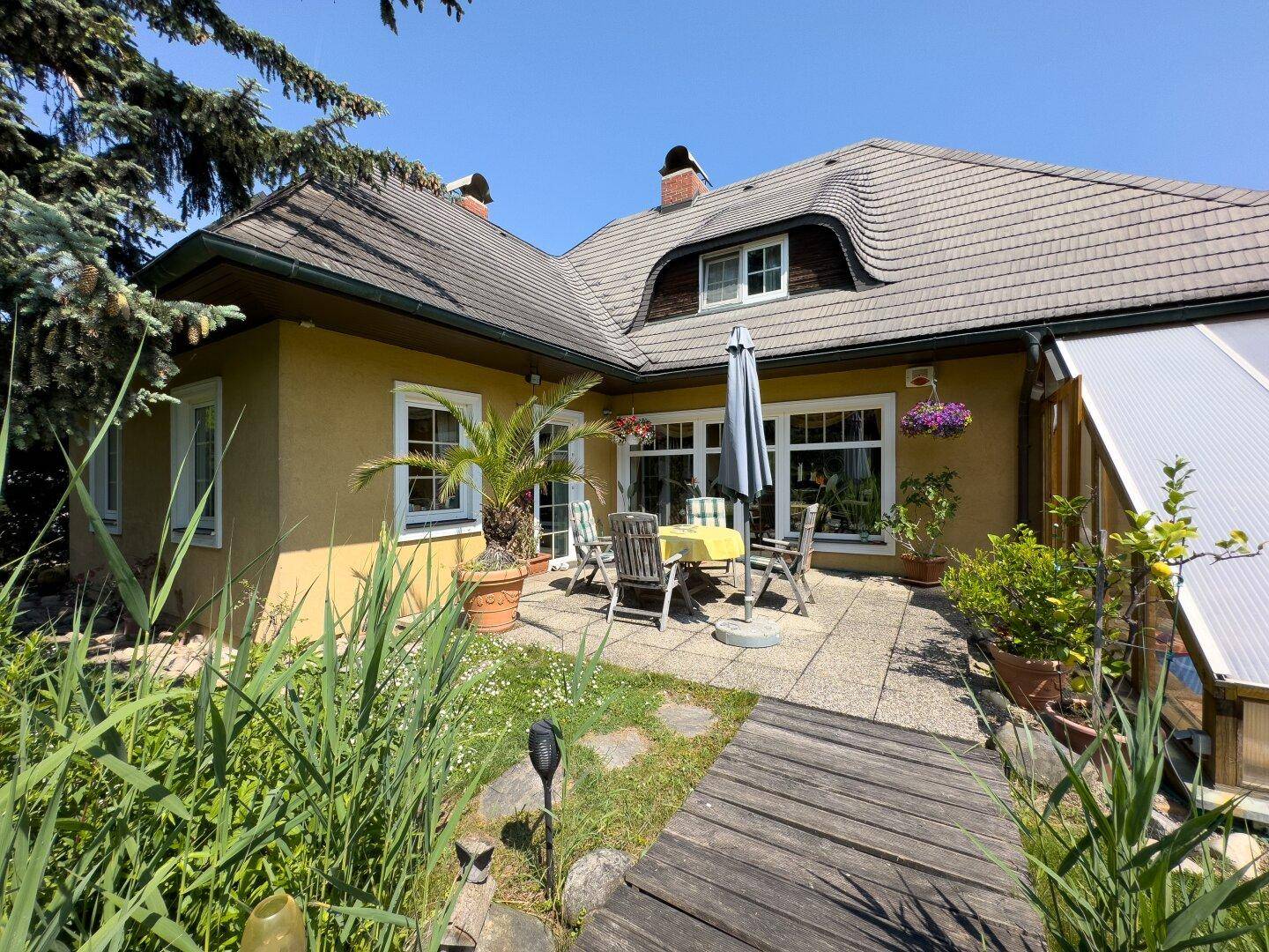 Sonnige Terrasse mit Gartenmöbeln und Zugang zum Haus, umgeben von üppigem Grün.
