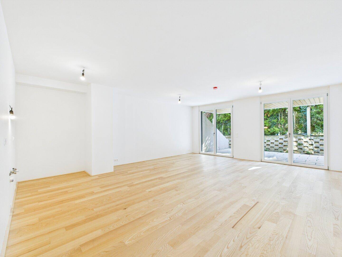 Heller, leerer Raum mit hellem Holzfußboden und großen Schiebetüren zur Terrasse mit Blick ins Grüne.