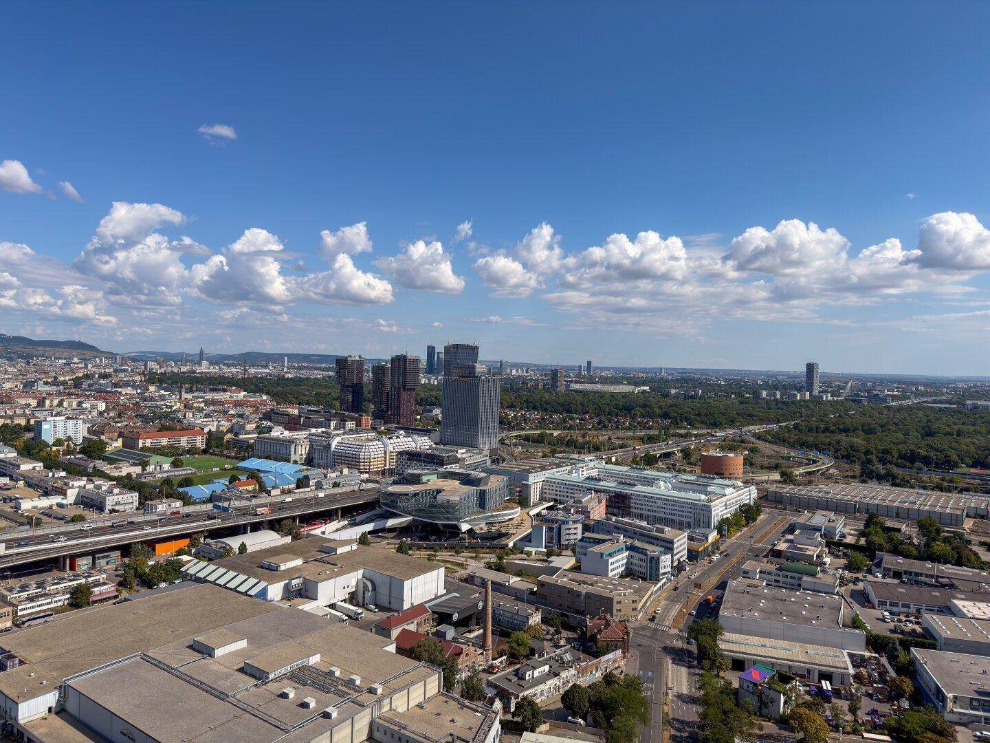 Panoramablick über die moderne Stadtlandschaft mit Hochhäusern, Grünflächen und Bergen am Horizont.