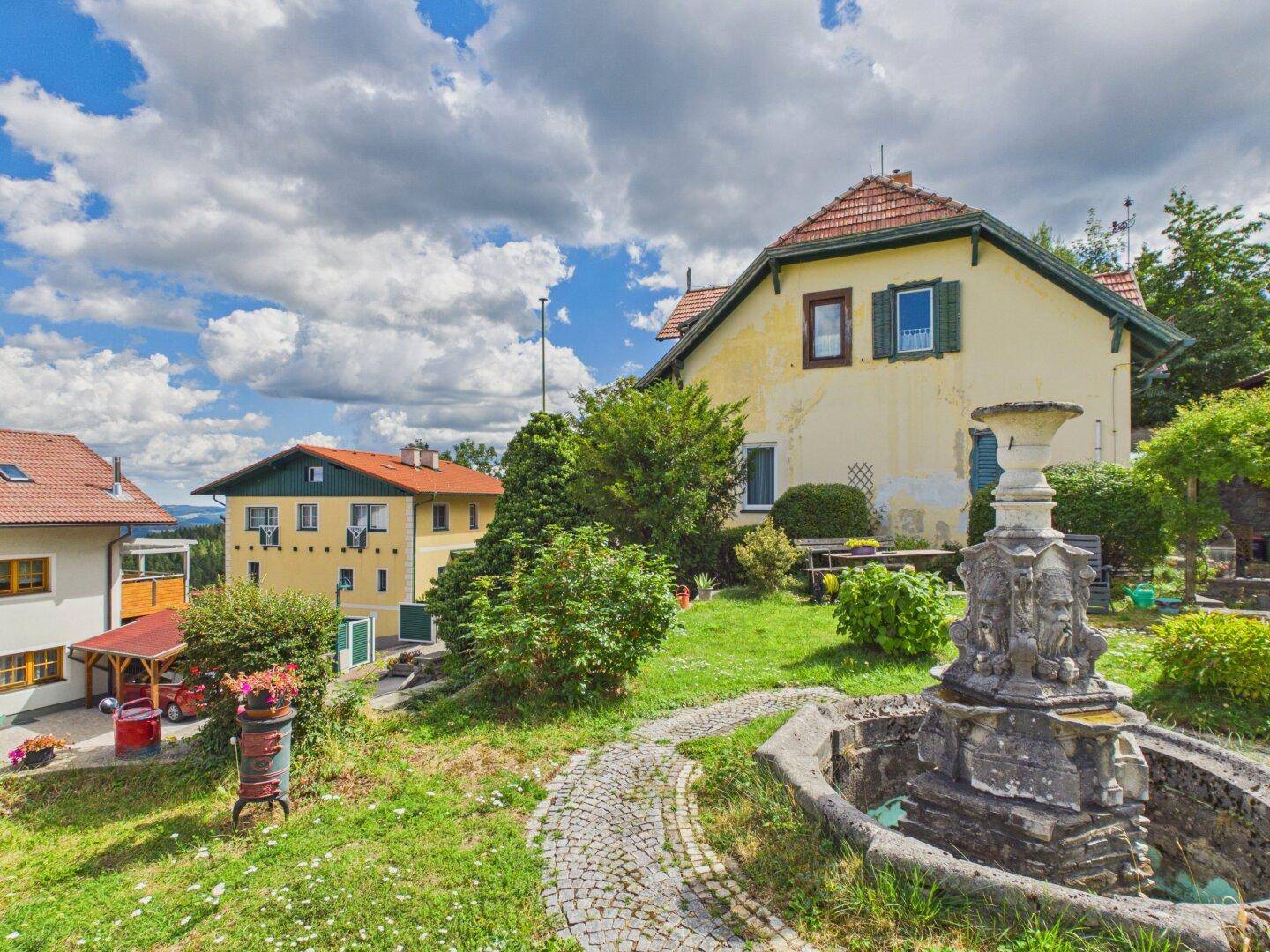 Malerischer Garten mit Steinbrunnen, gepflastertem Weg und Blick auf die umliegende Landschaft.