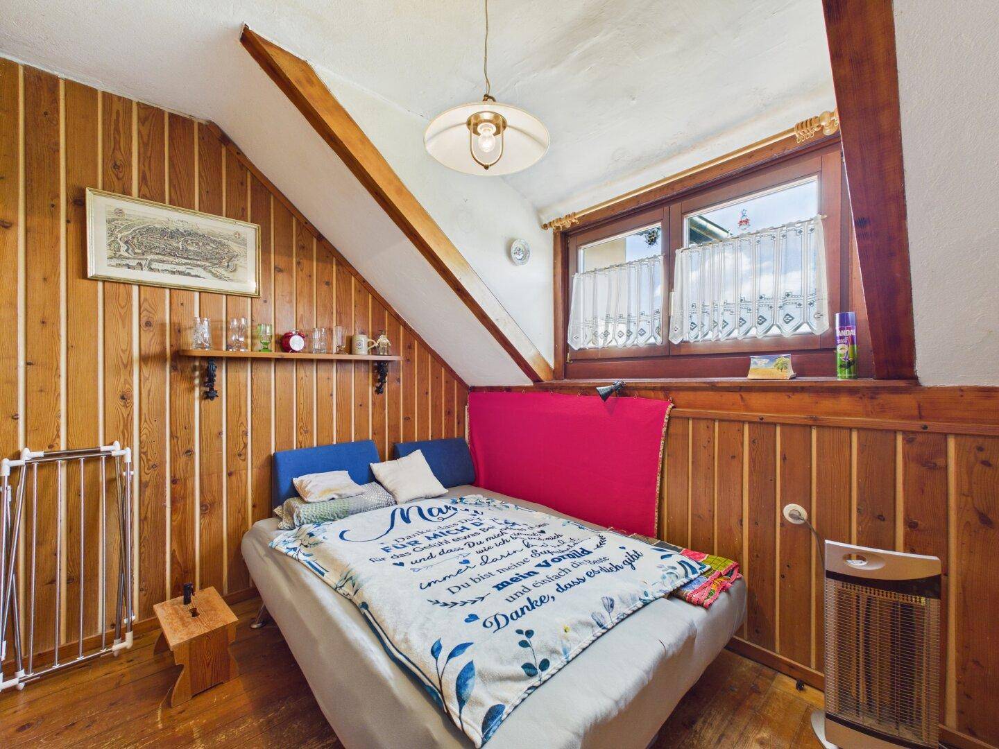 Schlafzimmer mit Holzvertäfelung, Holzboden und Dachschräge, unter einem Fenster.