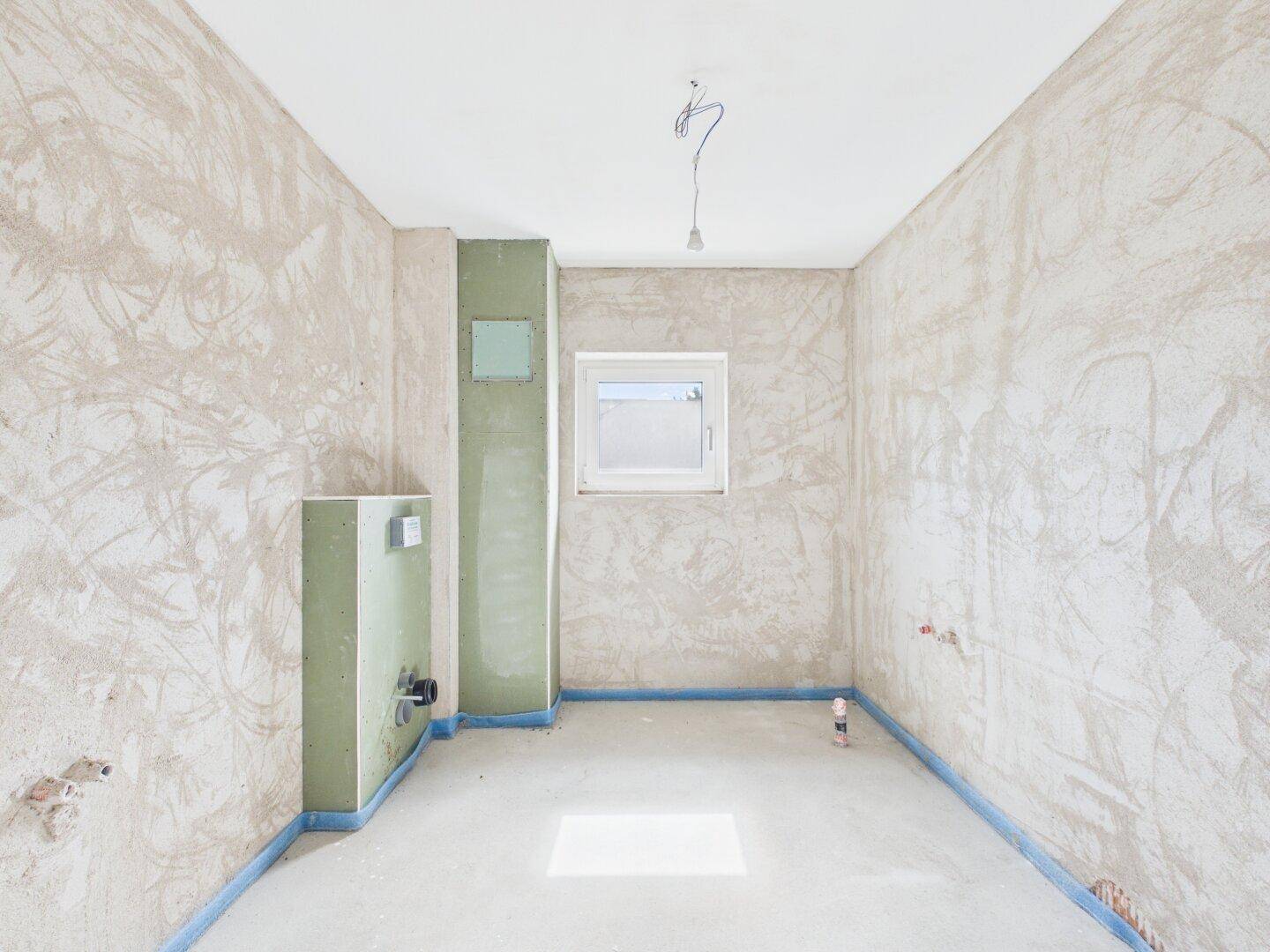 Unfertiges Badezimmer im Rohbau mit Fenster und vorbereiteten Installationen.