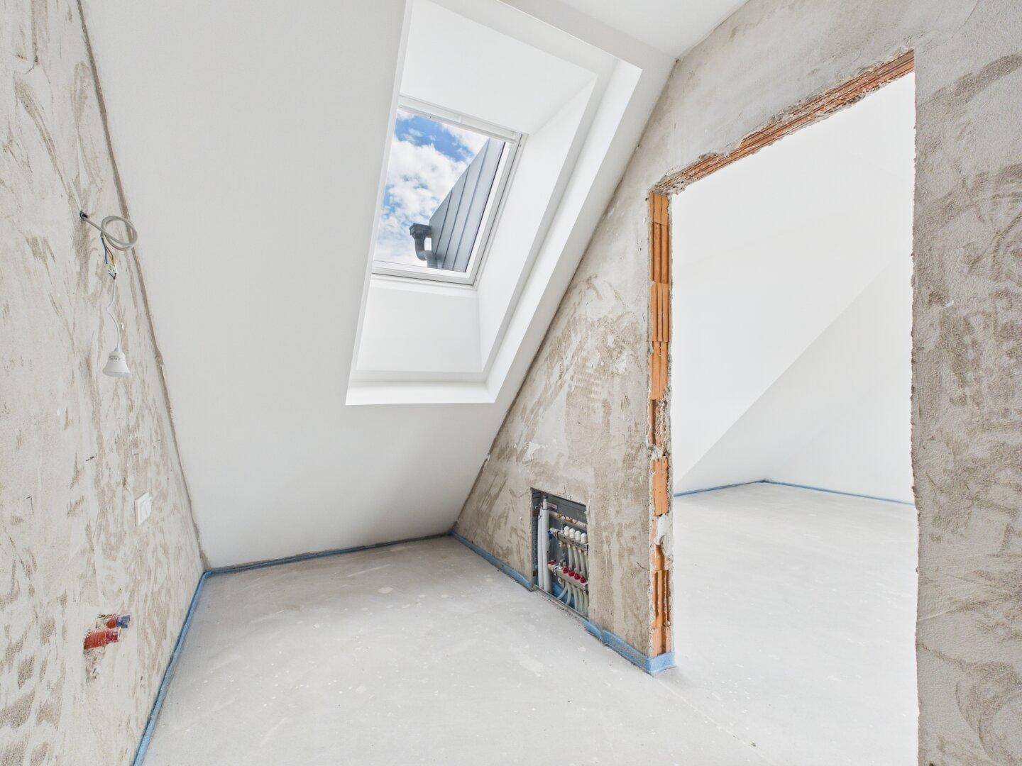 Unmöblierter Dachgeschossraum im Rohbau mit Dachschräge und Oberlicht.