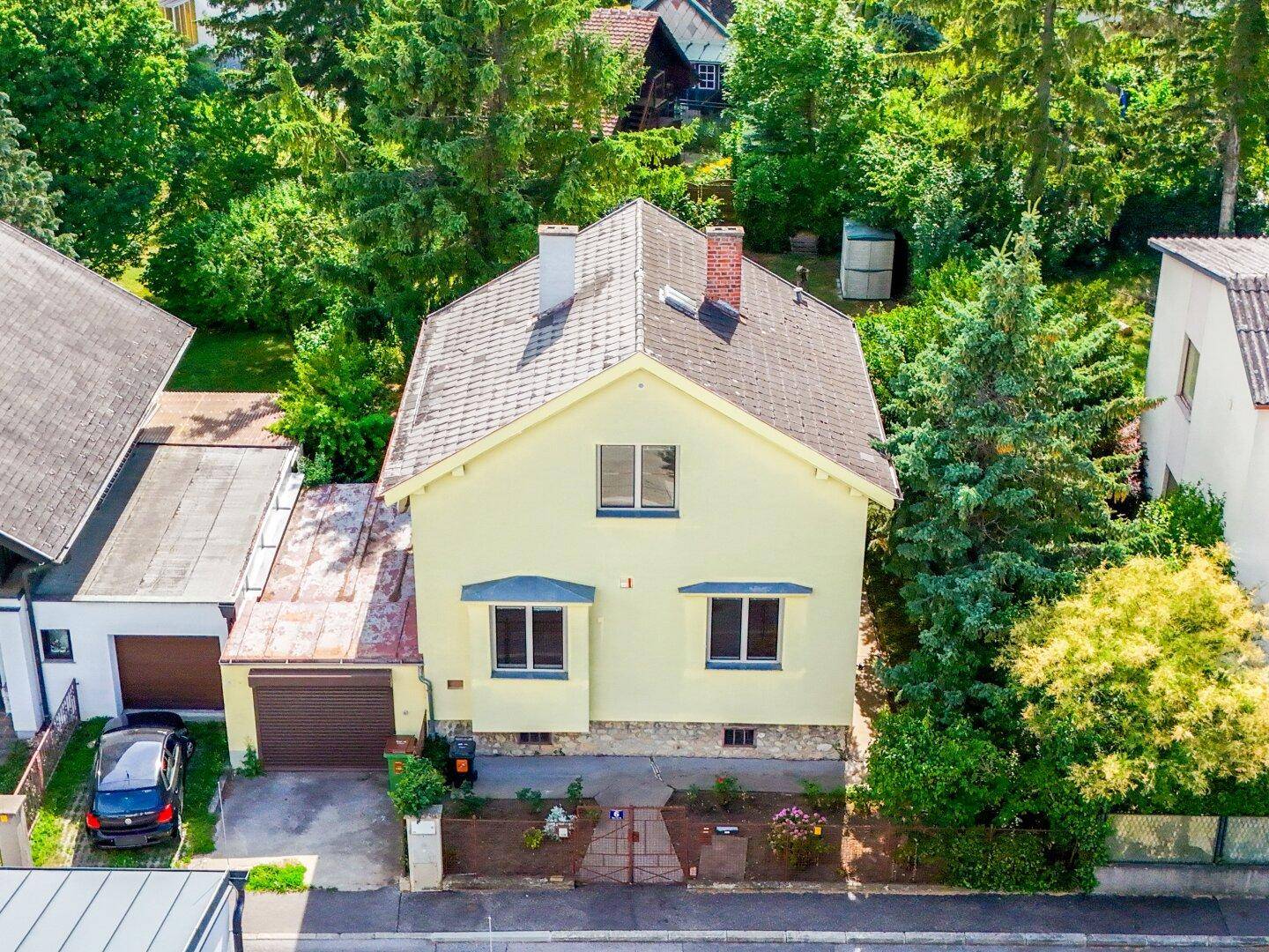 Luftaufnahme eines gelben Einfamilienhauses mit Satteldach, Garage und umgebender grüner Landschaft.