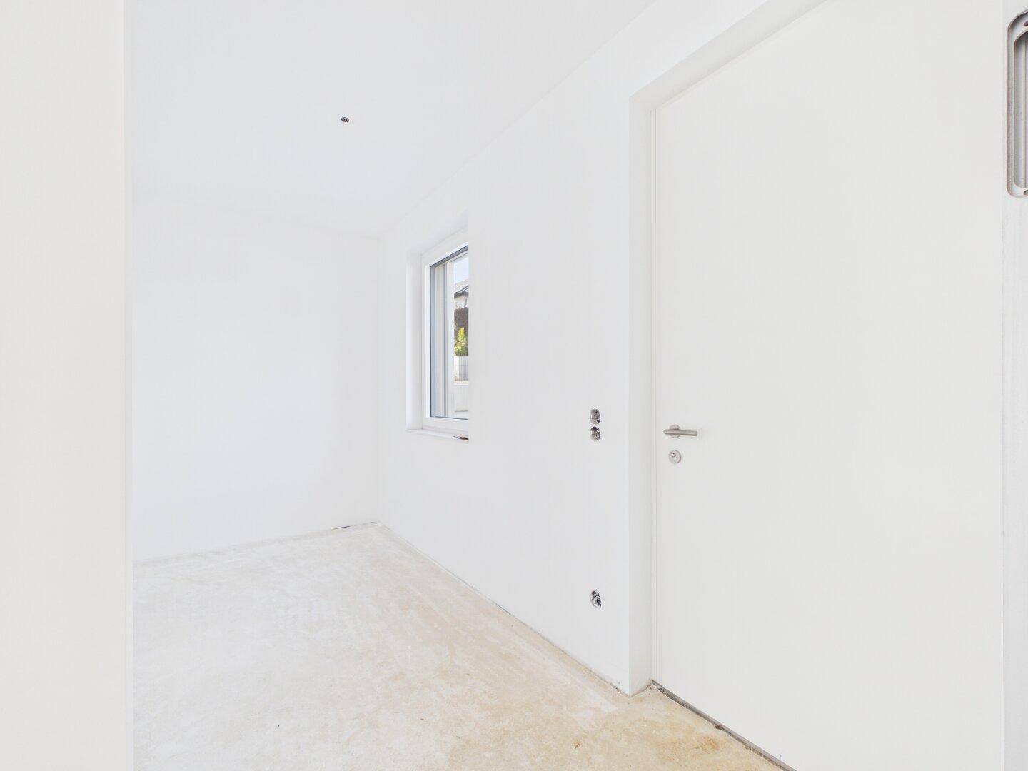Unmöblierter Raum im Rohbau mit weißer Tür und kleinem Fenster, bereit für den Ausbau.