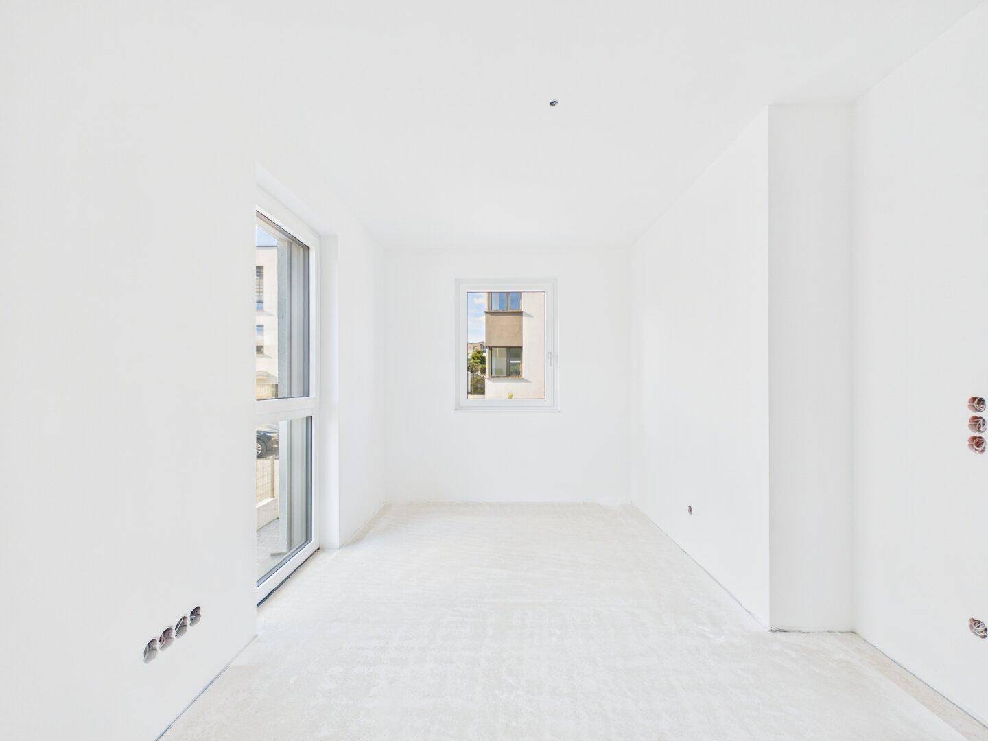 Helles Schlafzimmer im Rohbau mit großem Fenster und Zugang zu einem möglichen Balkon.