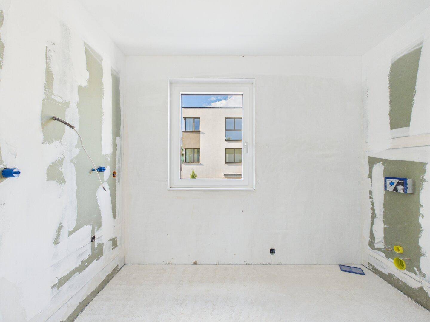 Raum im Rohbau mit einem Fenster und vorbereiteten Anschlüssen, bereit für den Innenausbau.