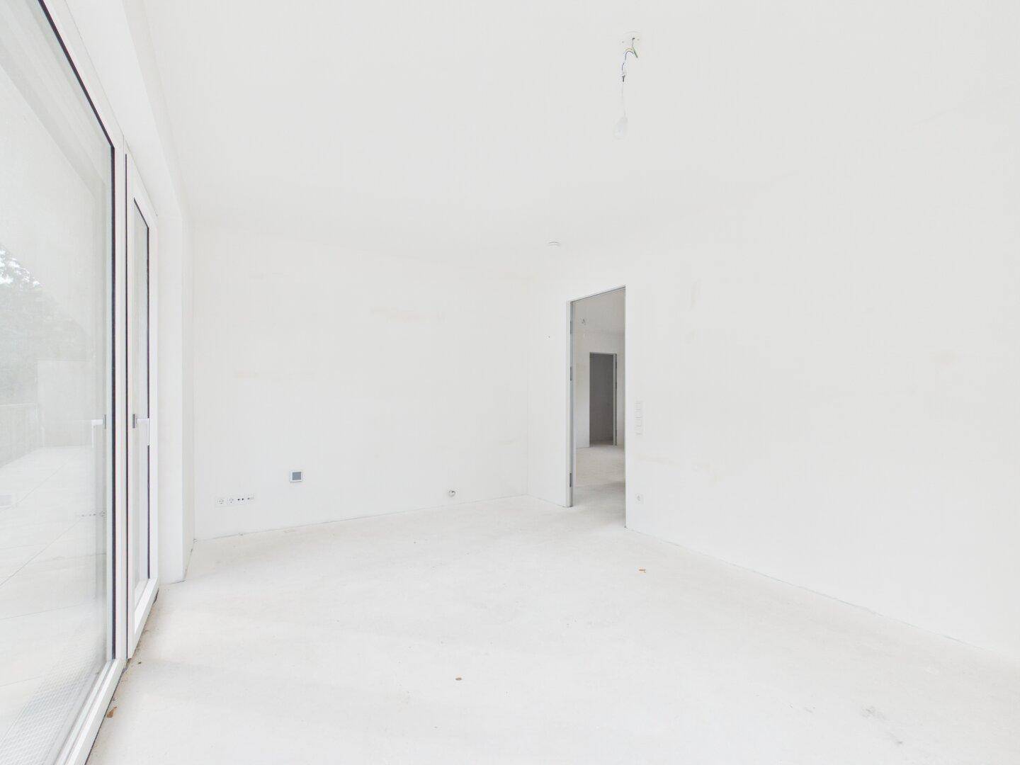 Leerer Raum im Rohbau mit einer Türöffnung und großen Fenstern, die den Blick nach draußen freigeben.