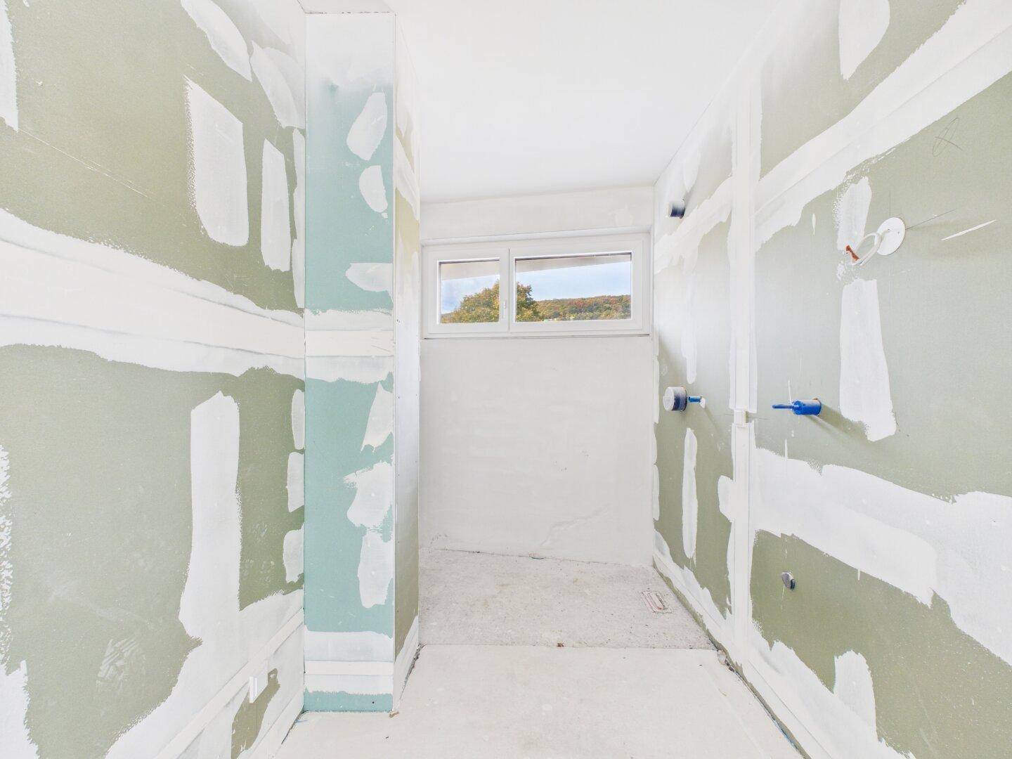 Unfertiges Badezimmer mit Fenster und vorbereiteten Anschlüssen für Sanitäranlagen.