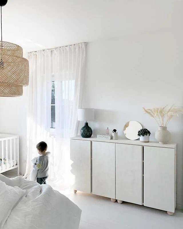Helles Schlafzimmer mit Babybett, weißem Holzboden und einem Kind am Fenster.