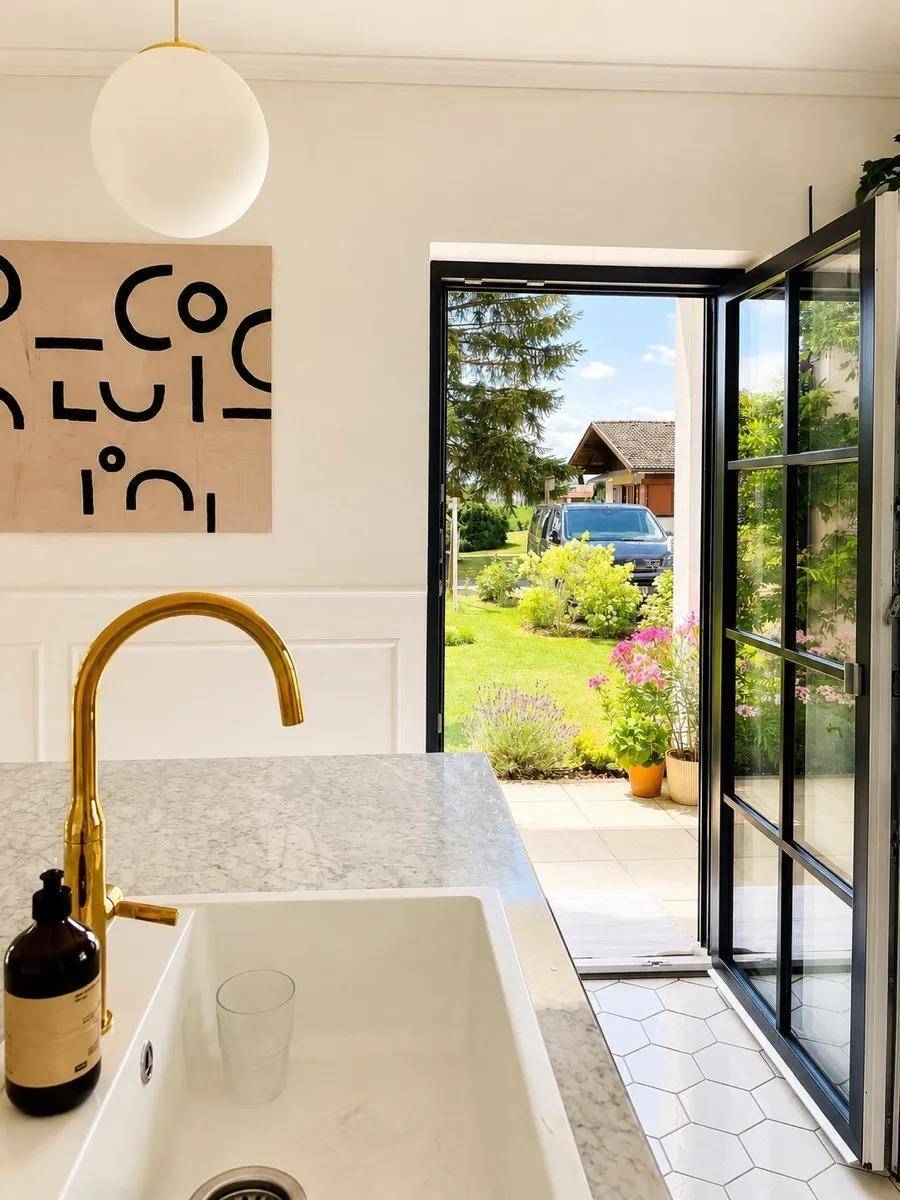 Moderne Küche mit Marmorarbeitsplatte, goldenem Wasserhahn und Blick in den grünen Garten.