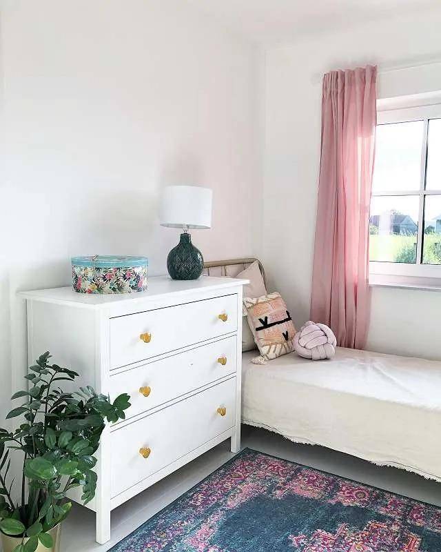 Kinderzimmer mit weißer Kommode, Einzelbett und rosa Vorhängen.