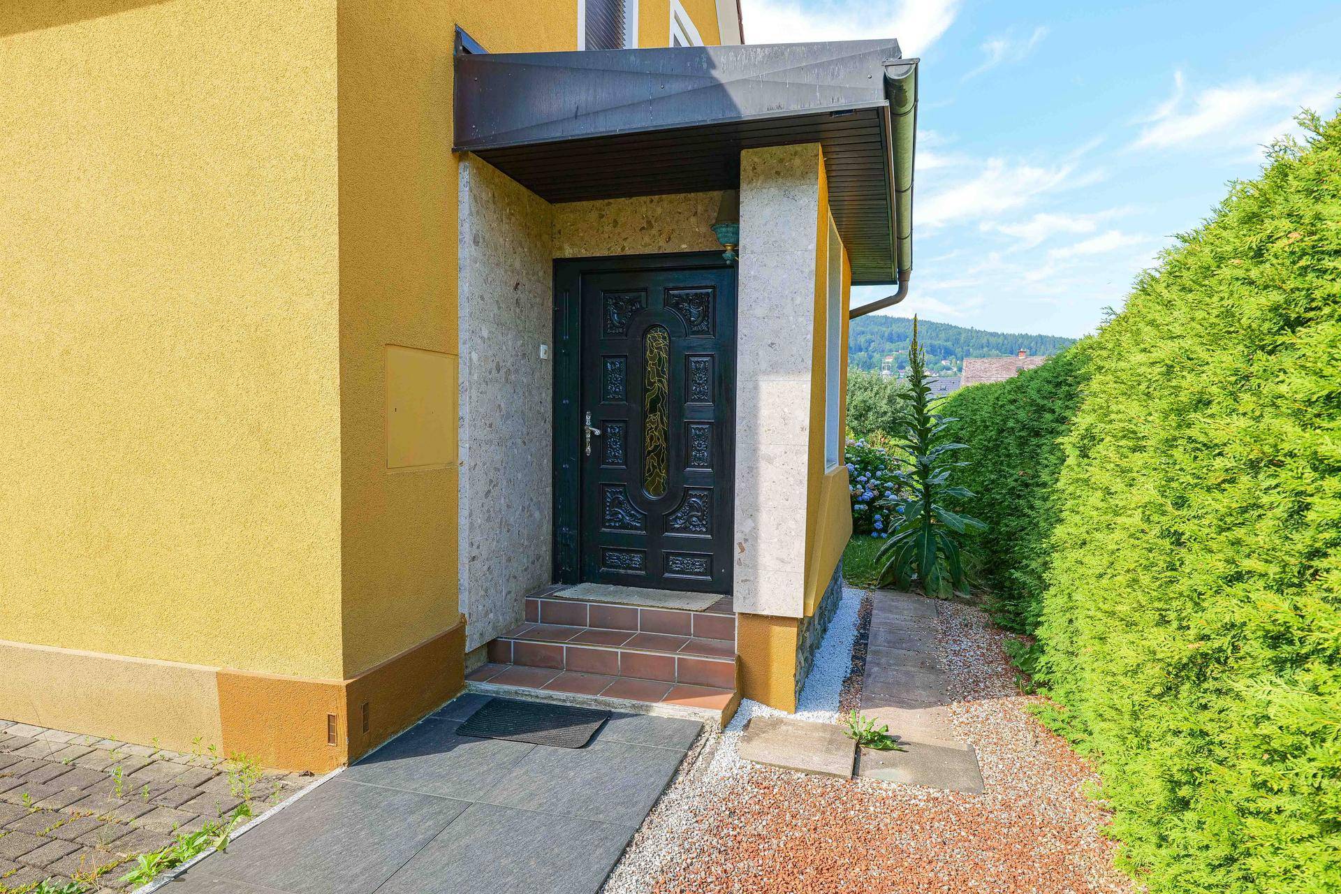 Überdachter Hauseingang mit dunkler Holztür und kleinen Kiesbeeten, umgeben von einer hohen Hecke.