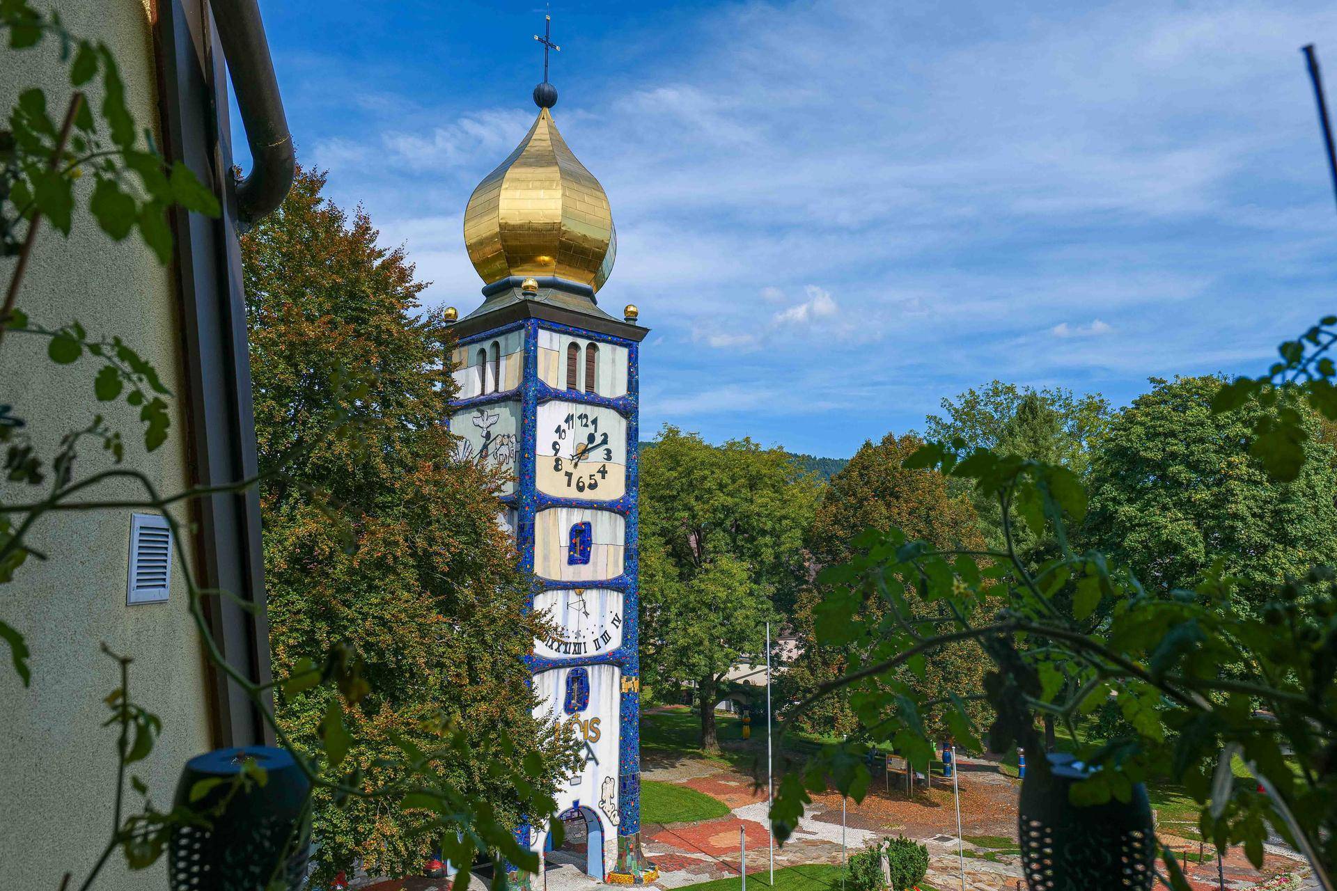 Nahaufnahme des historischen Kirchturms mit Uhr und goldener Kuppel, umgeben von Bäumen.