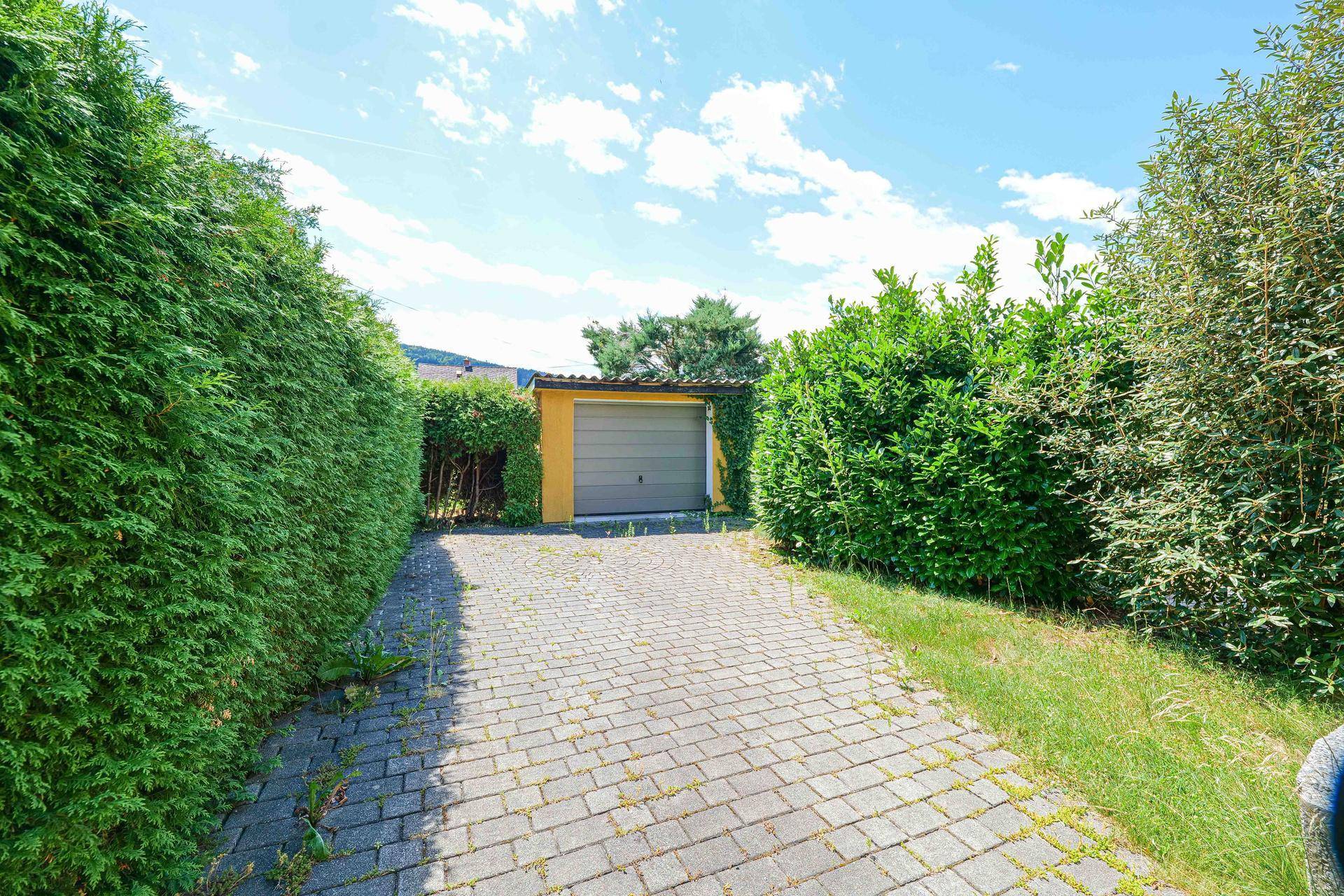 Gepflasterte Einfahrt führt zu einer freistehenden Garage, umgeben von hohen Hecken und Bäumen.