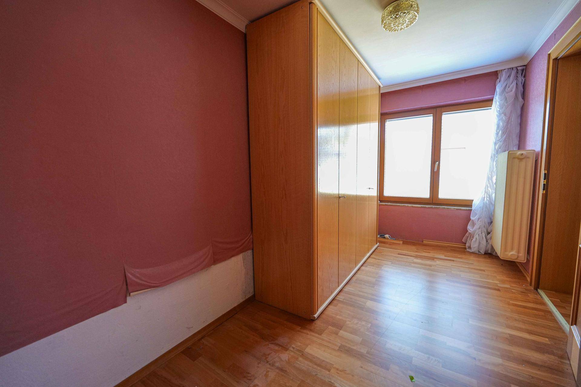 Schlafzimmer mit Holzboden, rosa Wänden und großem Kleiderschrank, gut beleuchtet durch ein Fenster.