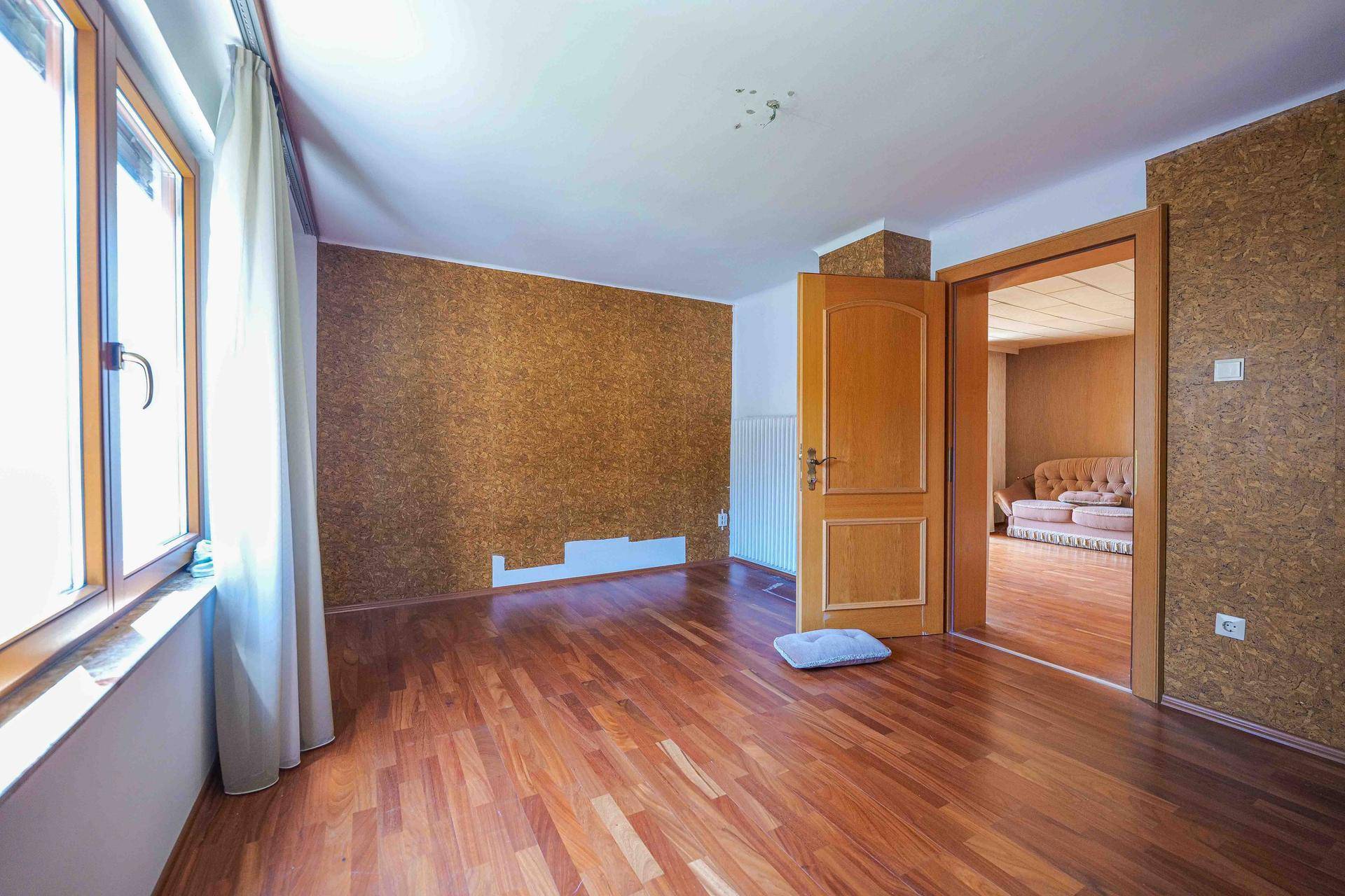 Helles Zimmer mit Holzboden und Korktapete, gut beleuchtet durch ein großes Fenster.