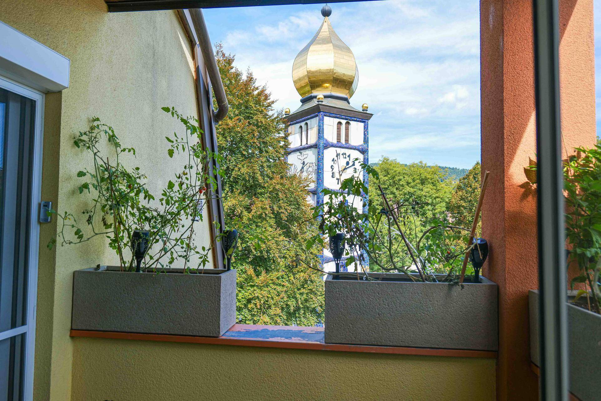 Blick vom Balkon auf einen markanten Kirchturm mit goldener Kuppel und üppigem Grün.