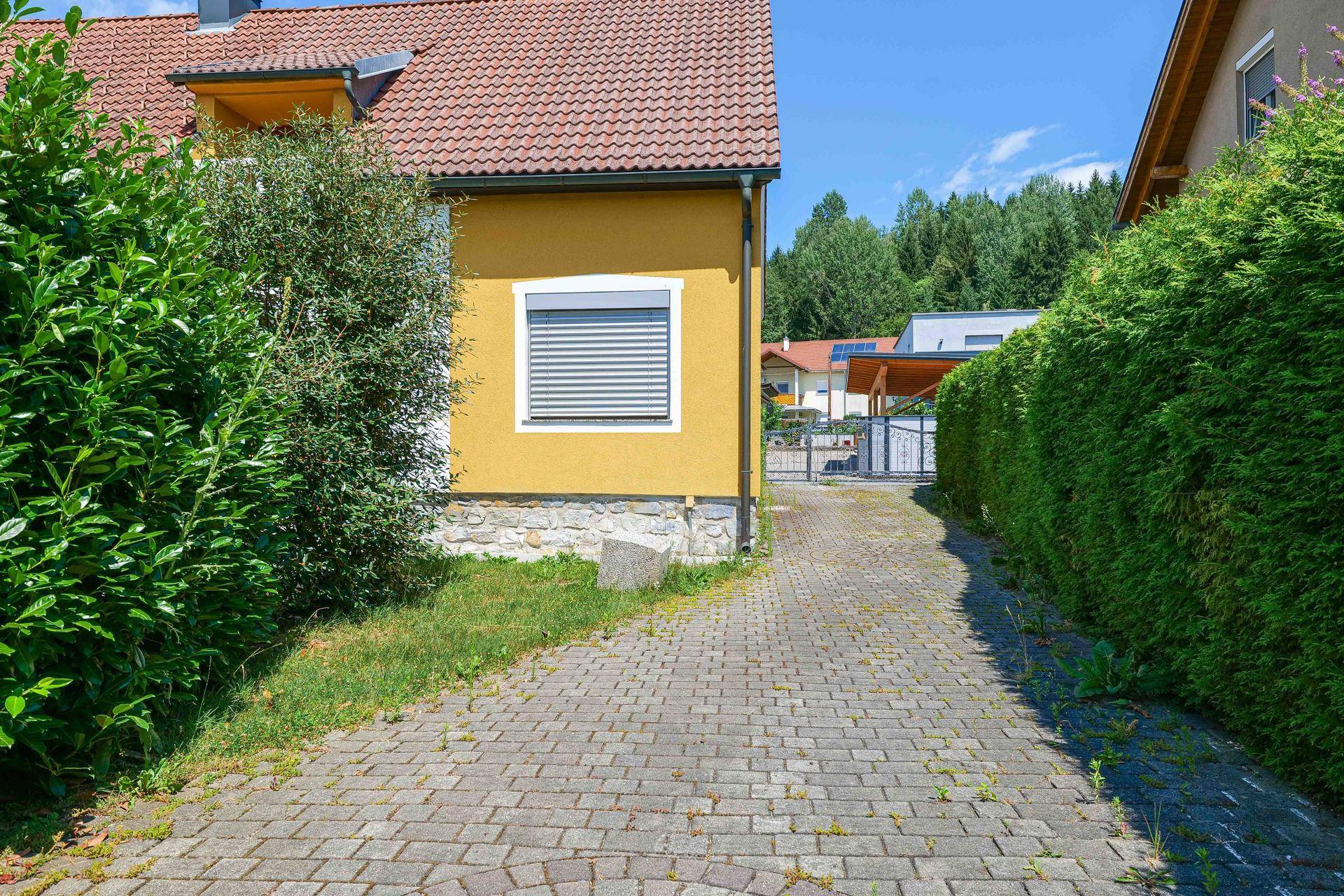 Seitenansicht des gelben Hauses mit gepflasterter Einfahrt und hoher, gepflegter Hecke unter blauem Himmel.