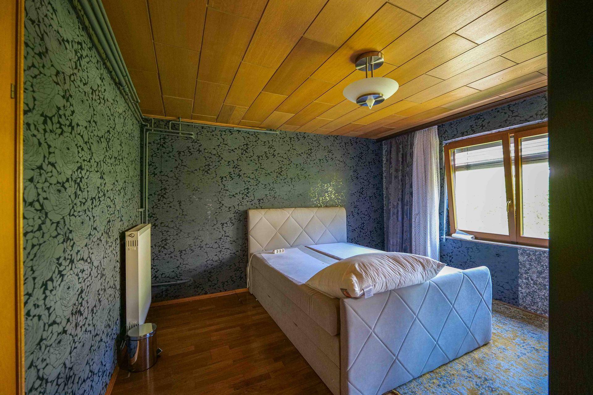 Schlafzimmer mit Doppelbett, Holzboden und dunkler gemusterter Tapete, gut beleuchtet durch ein Fenster.