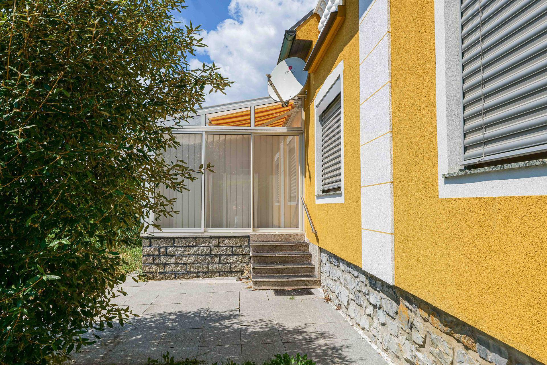 Seitlicher Zugang zum Wintergarten mit Steintreppe und Satellitenschüssel an der gelben Hauswand.