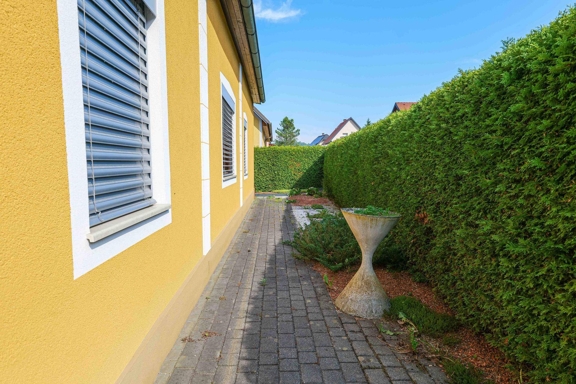 Schmaler gepflasterter Weg entlang der Hauswand mit hoher Hecke und dekorativer Steinvase.