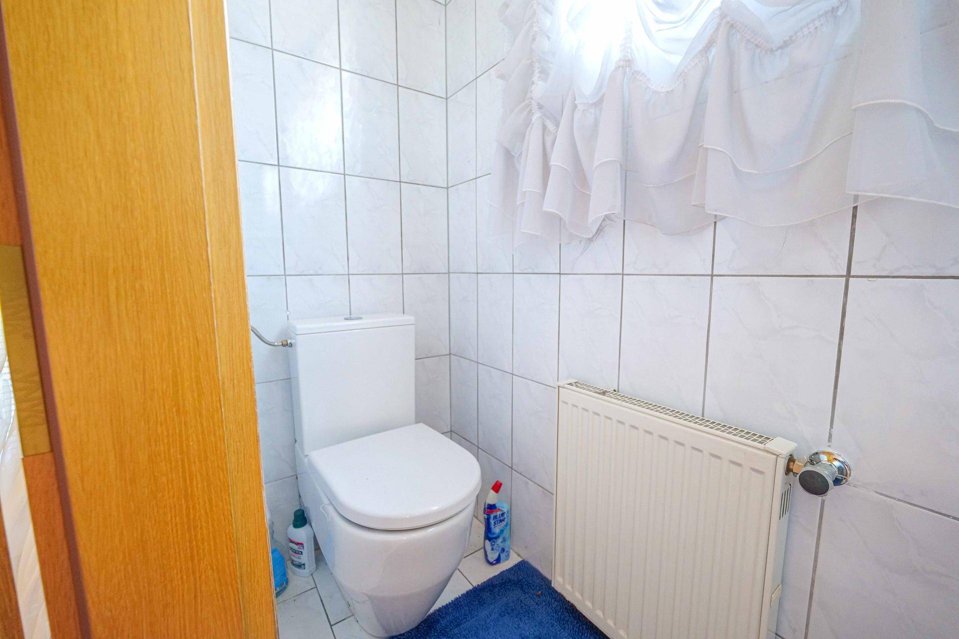 Kleines WC mit weißen Fliesen, Standard-Toilette und Heizkörper, beleuchtet durch ein Fenster.