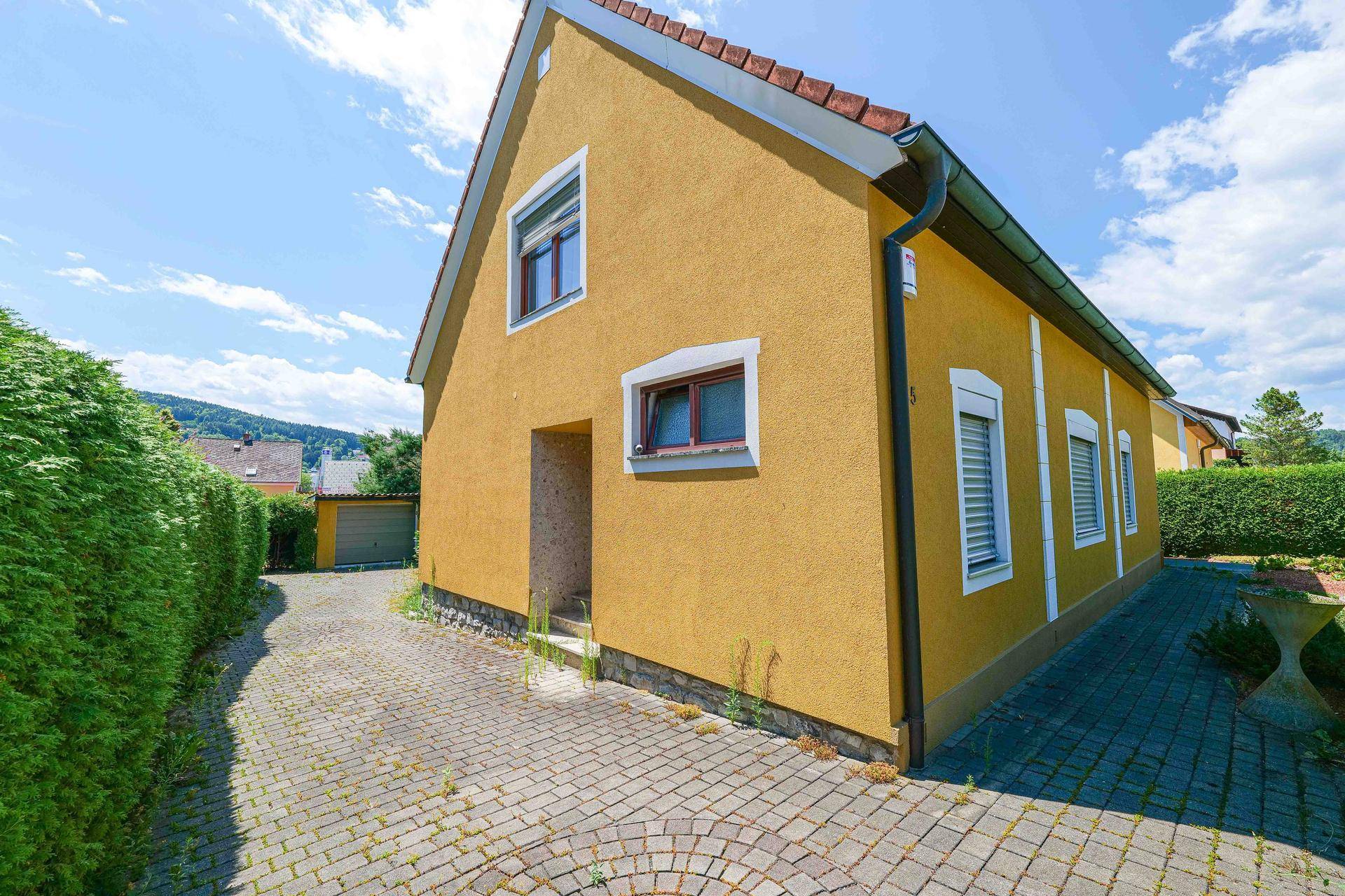 Seitenansicht des gelben Hauses mit gepflasterter Einfahrt, hoher Hecke und Garage im Hintergrund.
