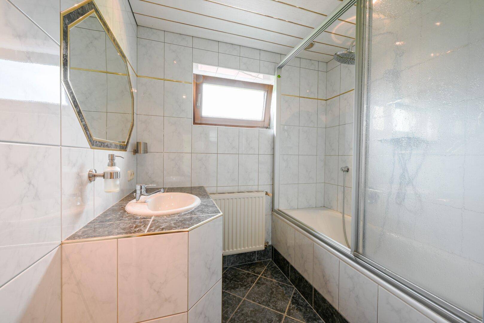 Badezimmer mit weißen Fliesen, Waschbecken und einer Badewanne mit Duschabtrennung.