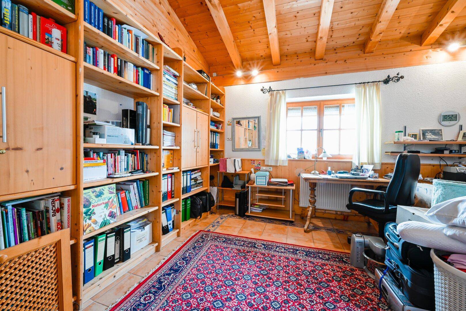 Gemütliches Büro mit Holzbalkendecke, großen Bücherregalen, Fliesenboden und Teppich.