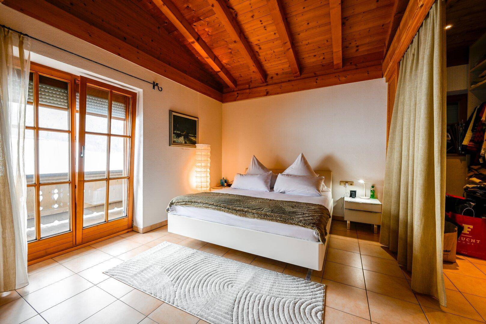 Helles Schlafzimmer mit Holzbalkendecke, Fliesenboden, großen Fenstern und Balkonzugang.