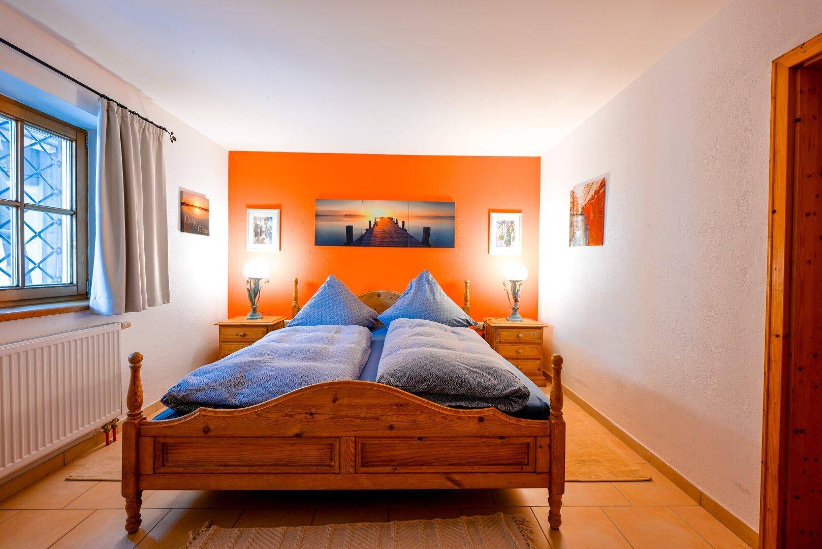 Schlafzimmer mit Holzbett, farbiger Akzentwand, Fliesenboden und Fenster.