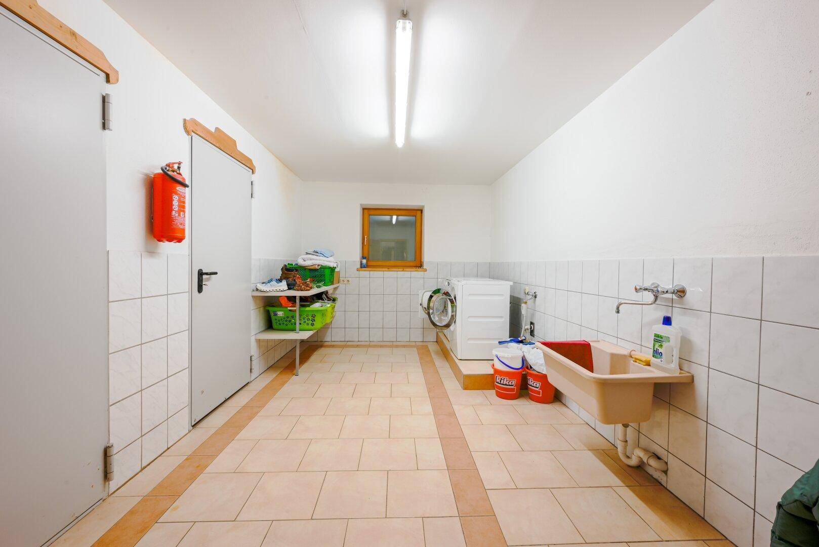 Praktischer Hauswirtschaftsraum mit Waschbecken, Waschmaschine, Fliesenboden und Fenster.