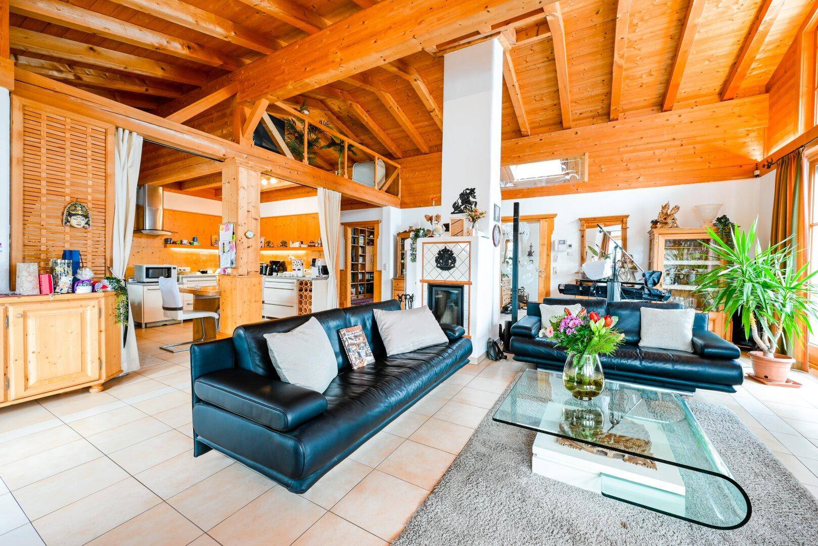 Großzügiges Wohnzimmer mit Ledersofas, Holzbalkendecke, Fliesenboden und offener Küche.