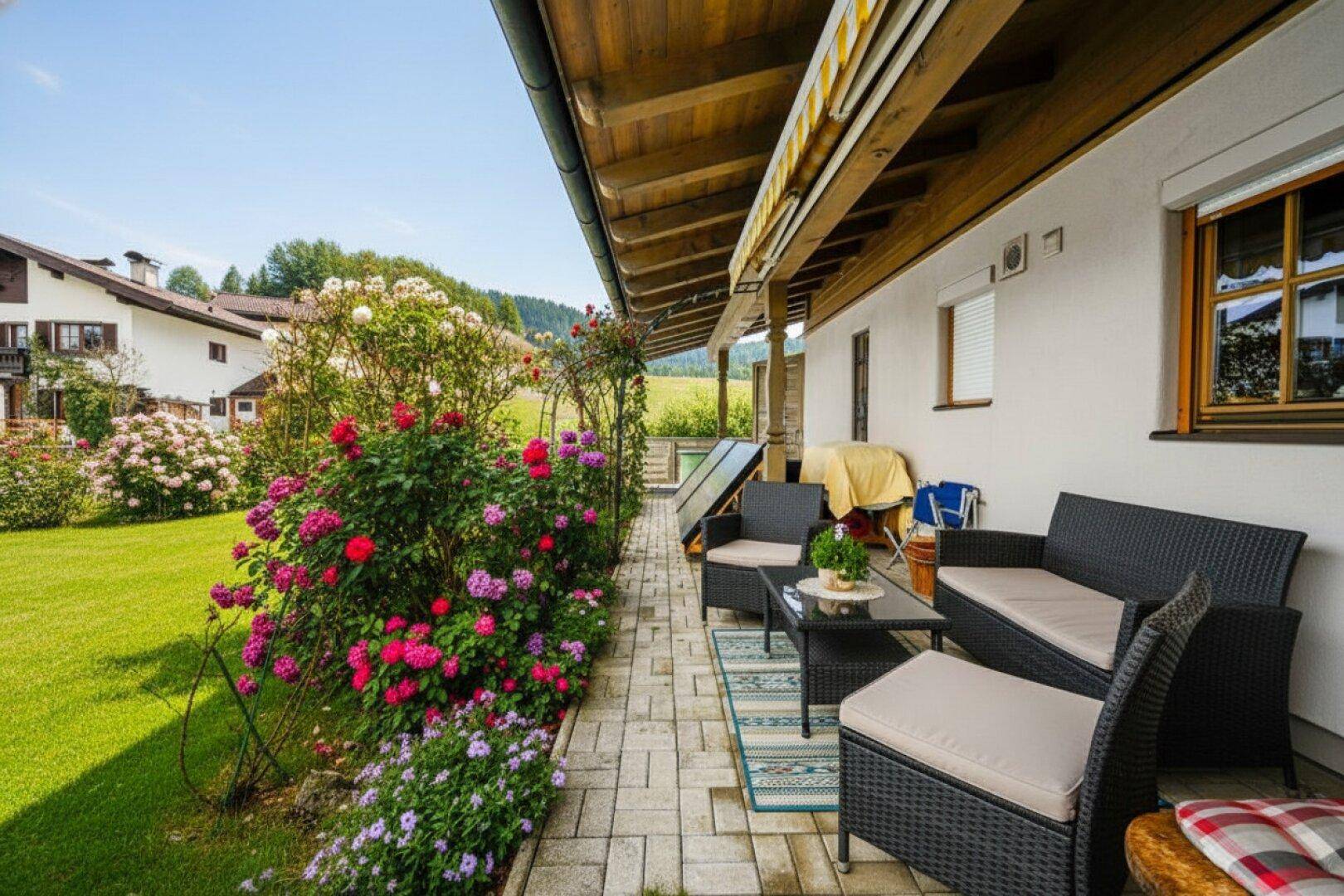 Sonniger Terrassenbereich mit Gartenmöbeln, Blumenbeeten, Rasen und Blick ins Grüne.