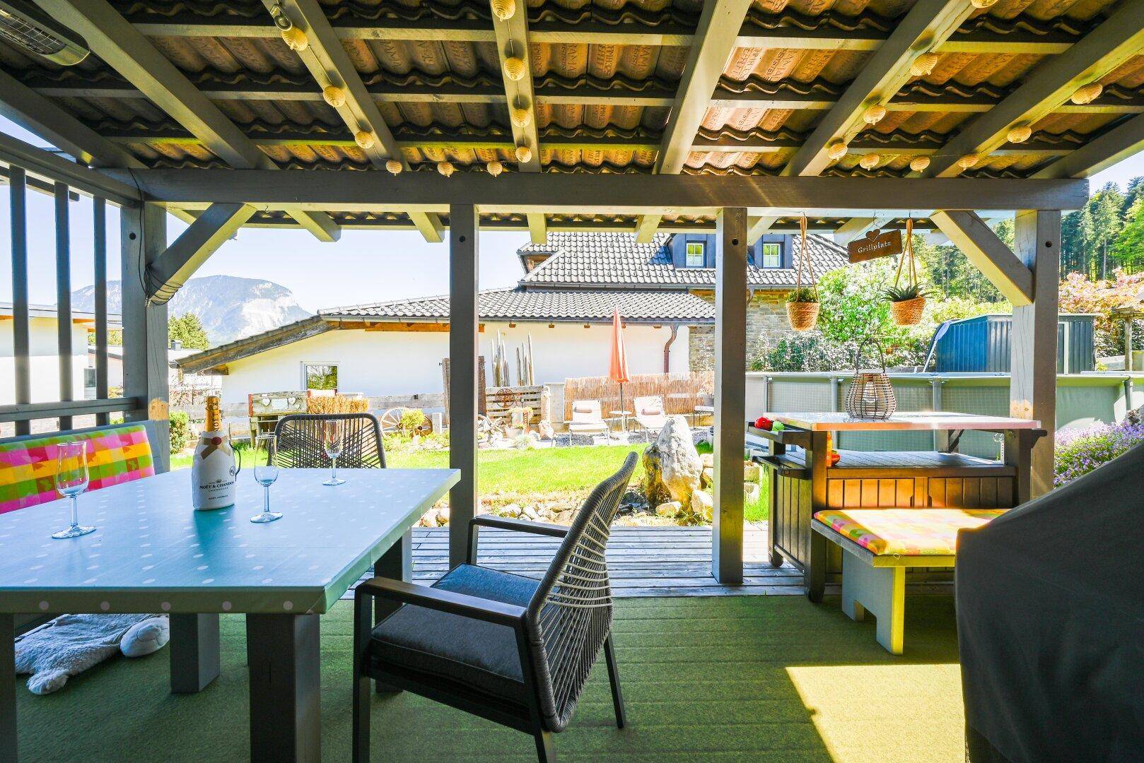 Überdachte Terrasse mit Esstisch und Stühlen, umgeben von Grün und Blick auf die Berge.