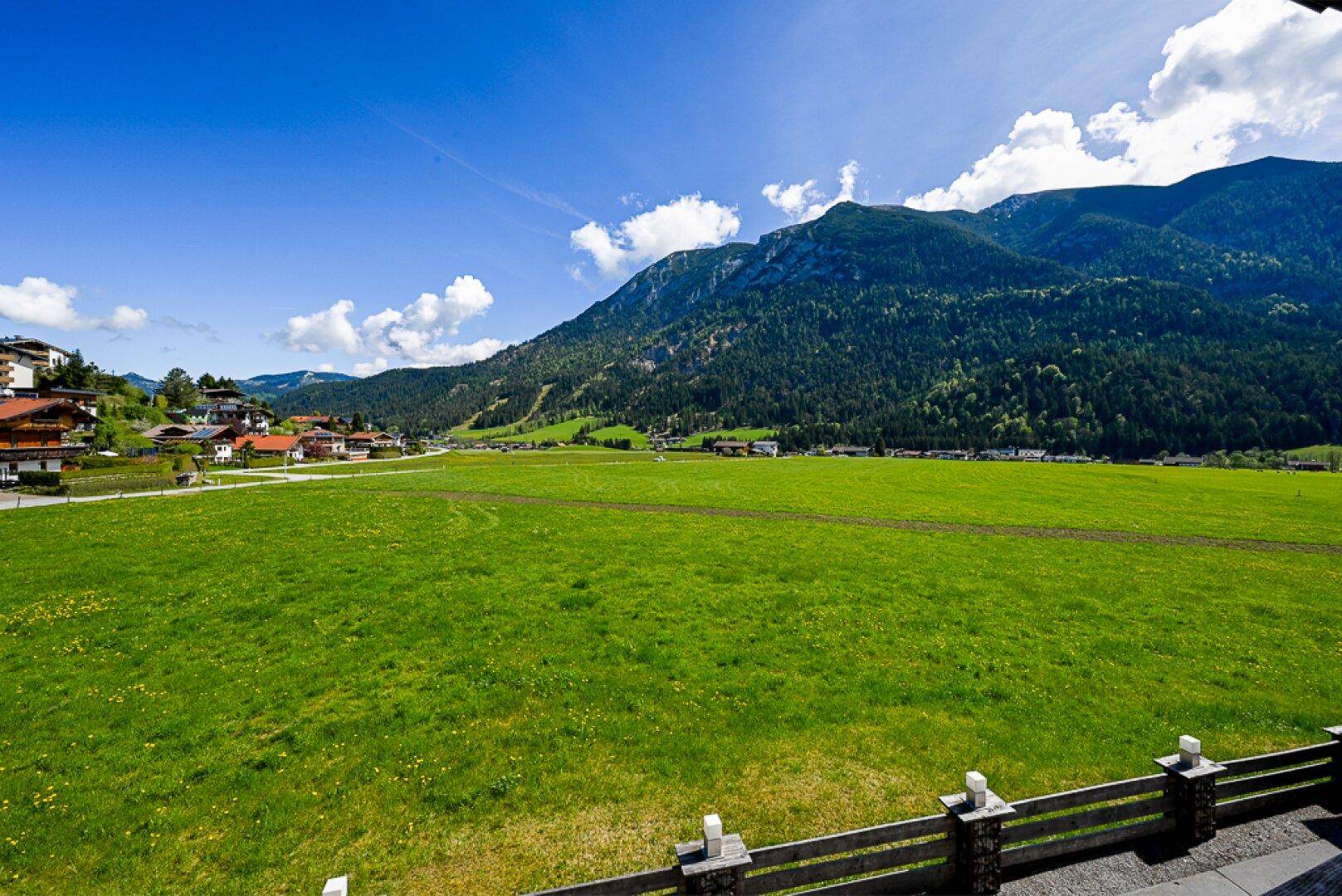 Weitläufige grüne Wiesen und Berge unter blauem Himmel, umgeben von ländlicher Bebauung.