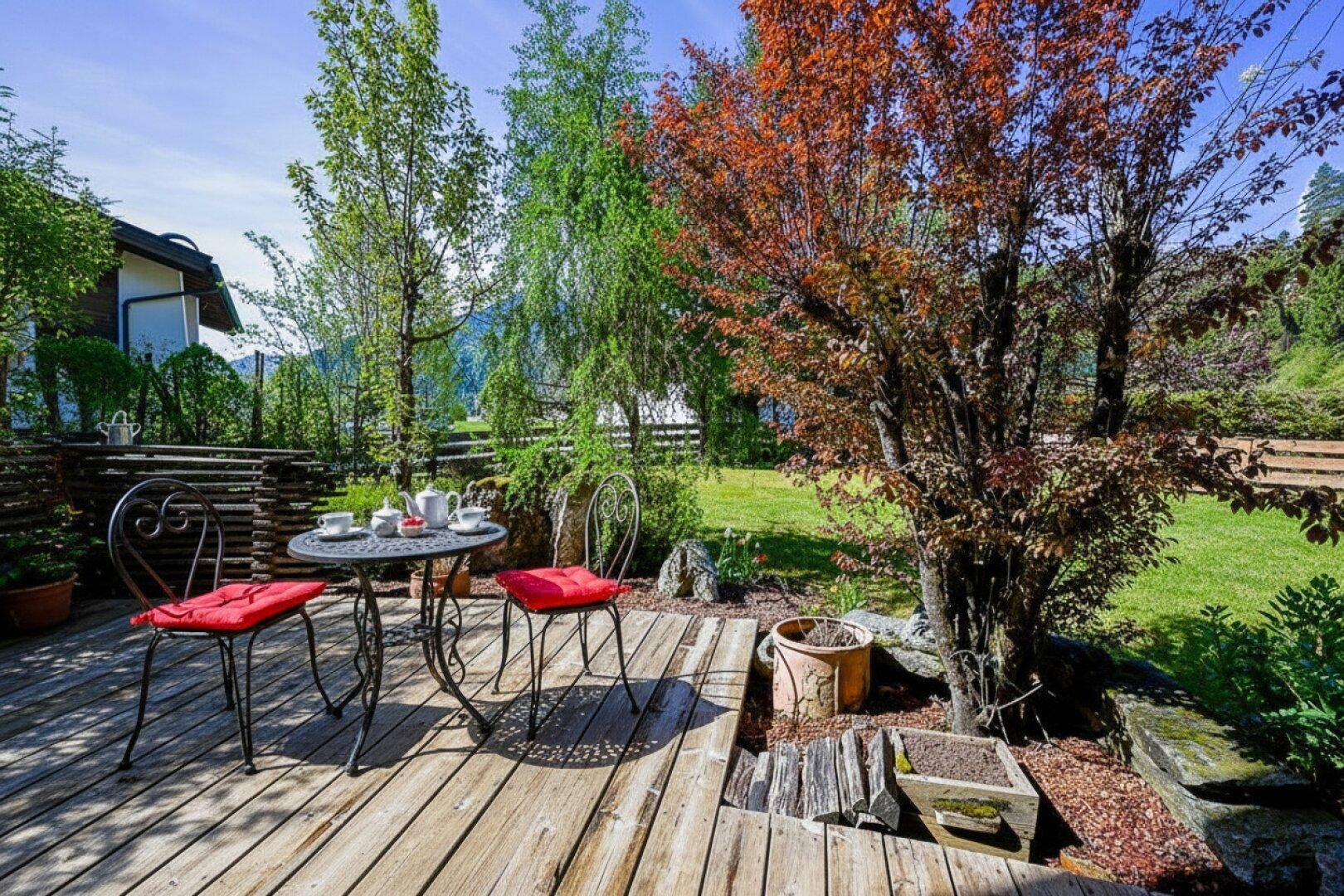 Gemütliche Sitzecke im Garten auf einer Holzterrasse, umgeben von Bäumen und Grünflächen.