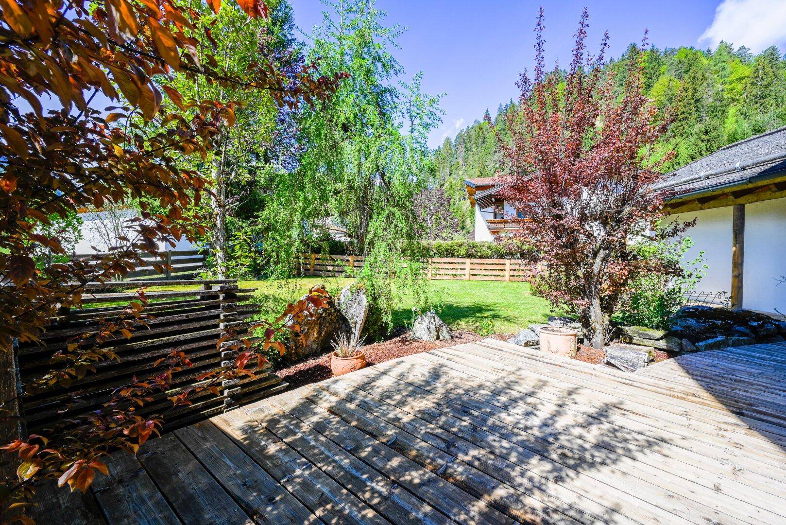 Holzterrasse im Garten mit Blick auf die grüne Landschaft und umliegende Bäume.